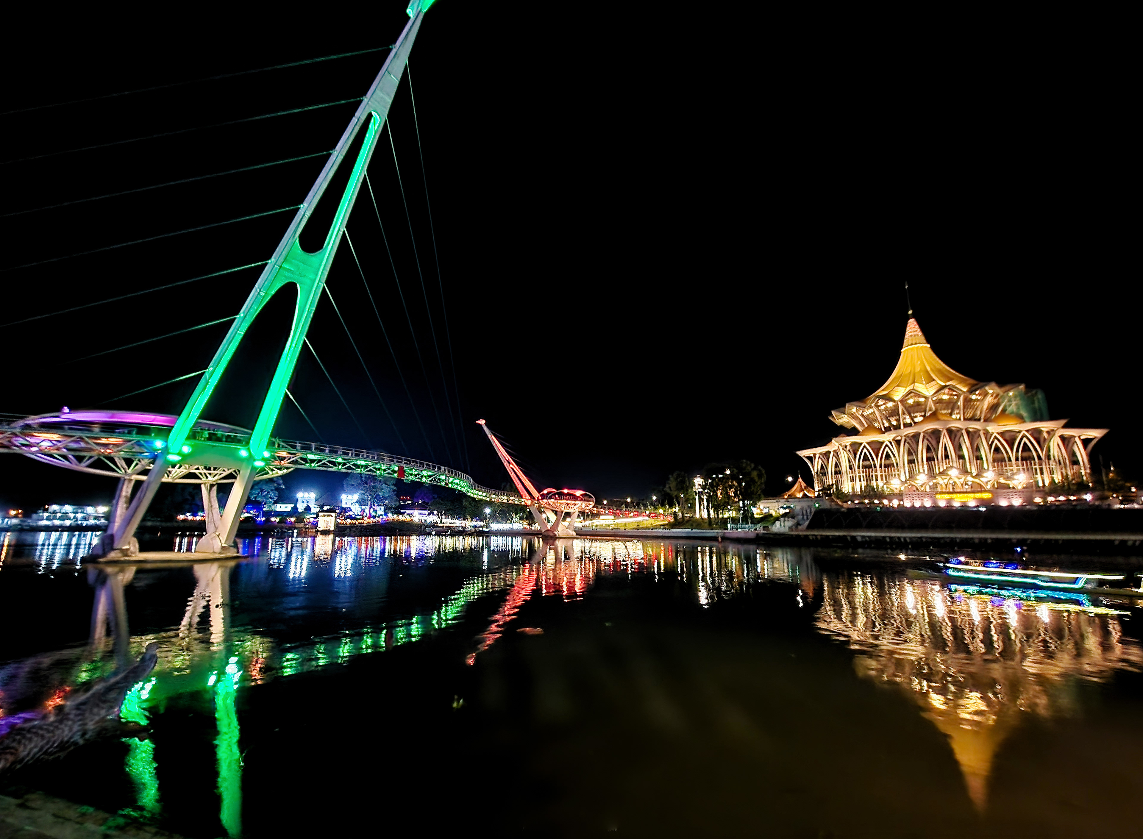 A vibrant nighttime cityscape featuring a modern suspension bridge illuminated in neon green and purple, juxtaposed against a grand, golden-lit building with a tiered roof, both casting vivid, colorful reflections across the dark, calm surface of a river.