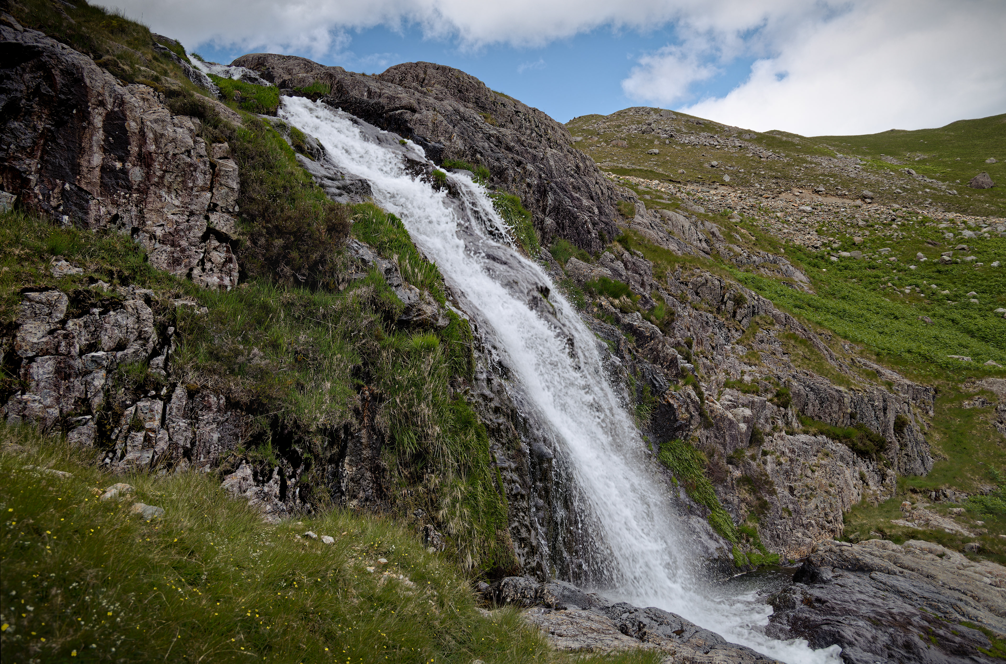 A fine-art landscape photograph showing a bright white waterfall rushing diagonally down a rugged, textured rock face dotted with vibrant green grass, backed by a rolling hill and a partly cloudy blue sky.