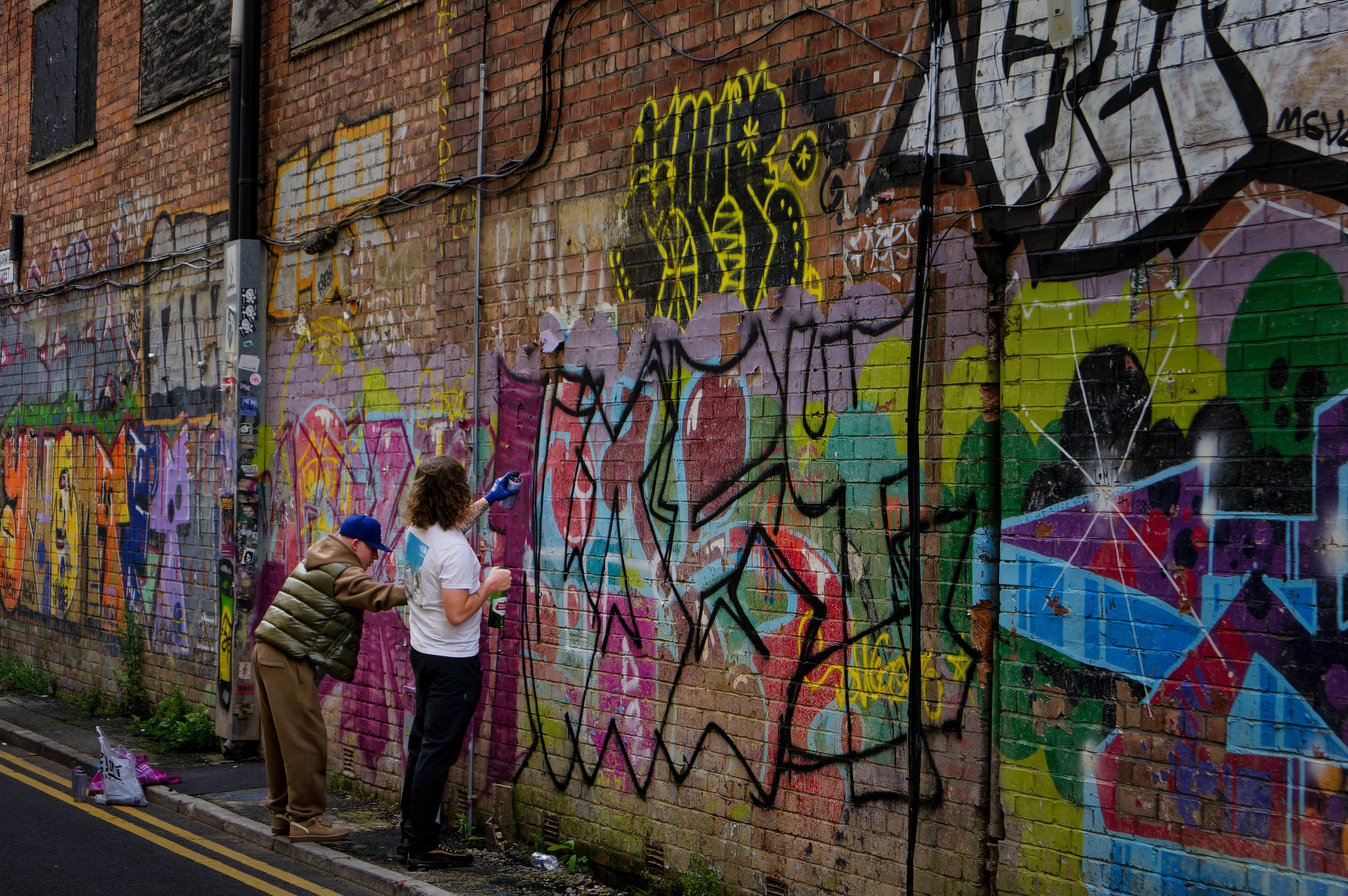 A vibrant fine-art street photograph capturing two figures in the act of spray-painting colorful graffiti onto a heavily textured, paint-layered brick wall in an urban alleyway, with discarded paint cans resting on the pavement below.