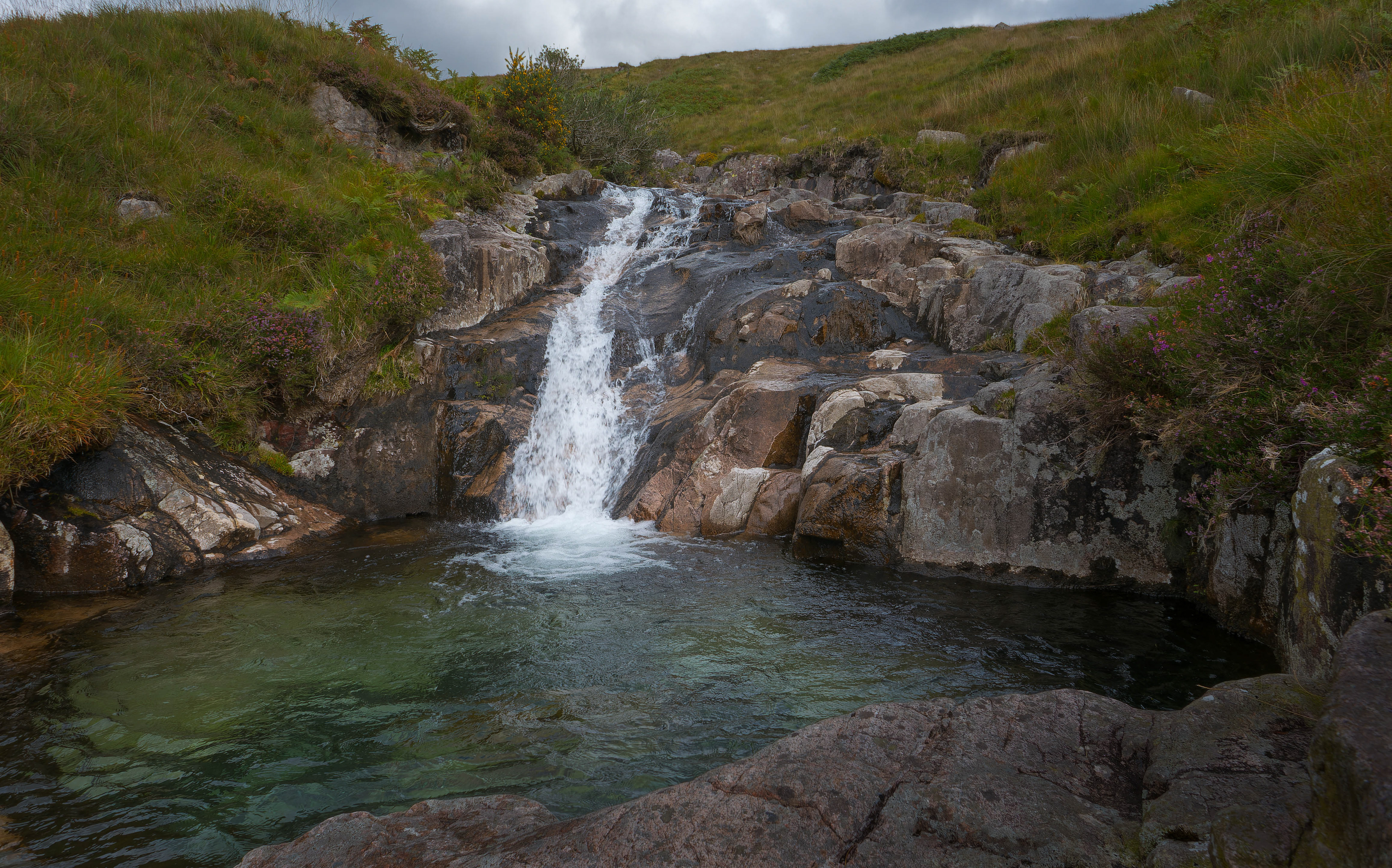 A fine-art landscape photograph showing a small waterfall cascading down a rugged, rocky incline into a remarkably clear, deep green pool, surrounded by wild moorland grasses and heather under a moody, overcast sky.