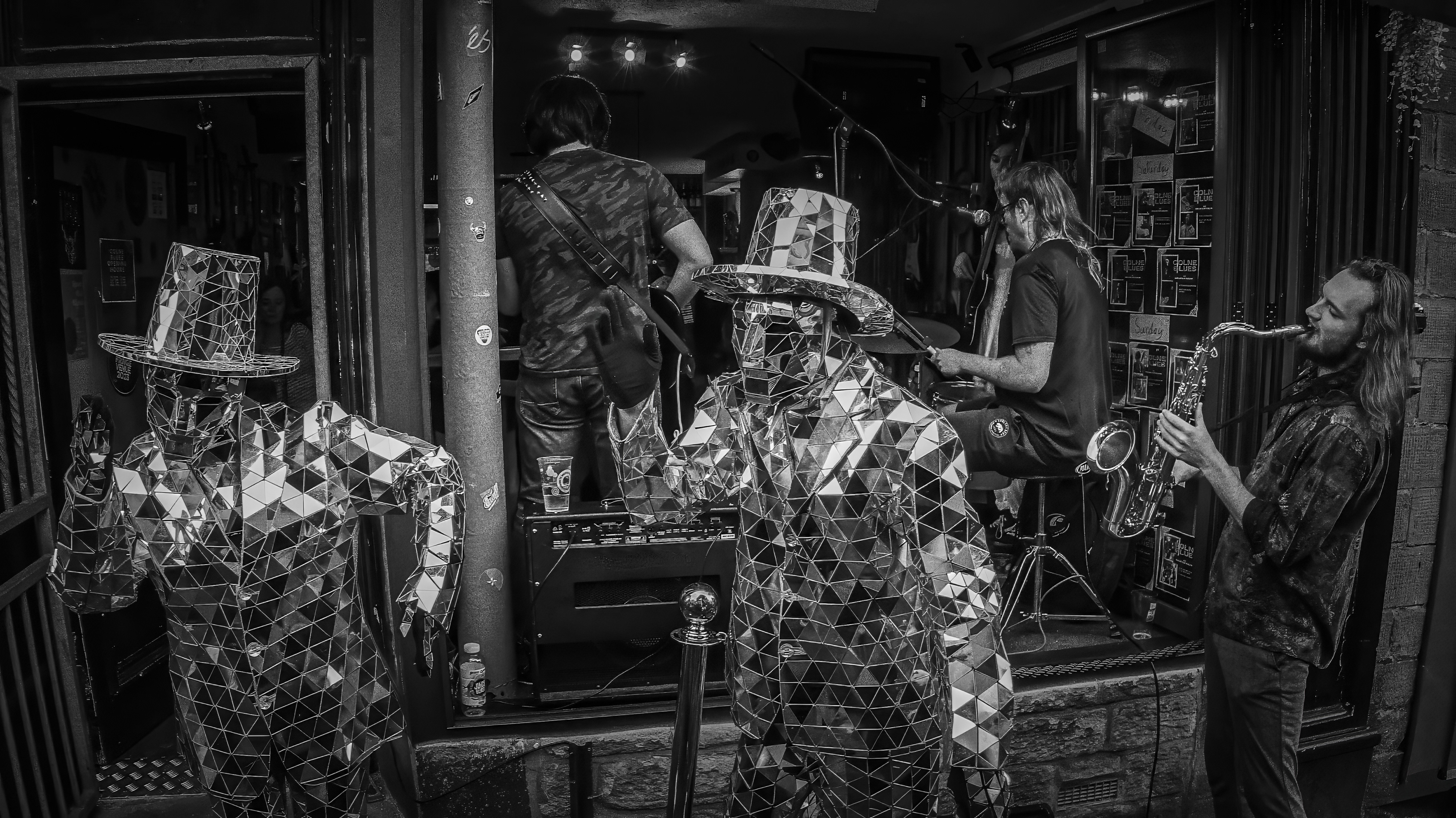 A high-contrast black and white photograph featuring two street performers in suits and top hats made entirely of small mirrors standing motionless outside a venue, juxtaposed against a live band with a saxophonist and drummer playing energetically in the background.