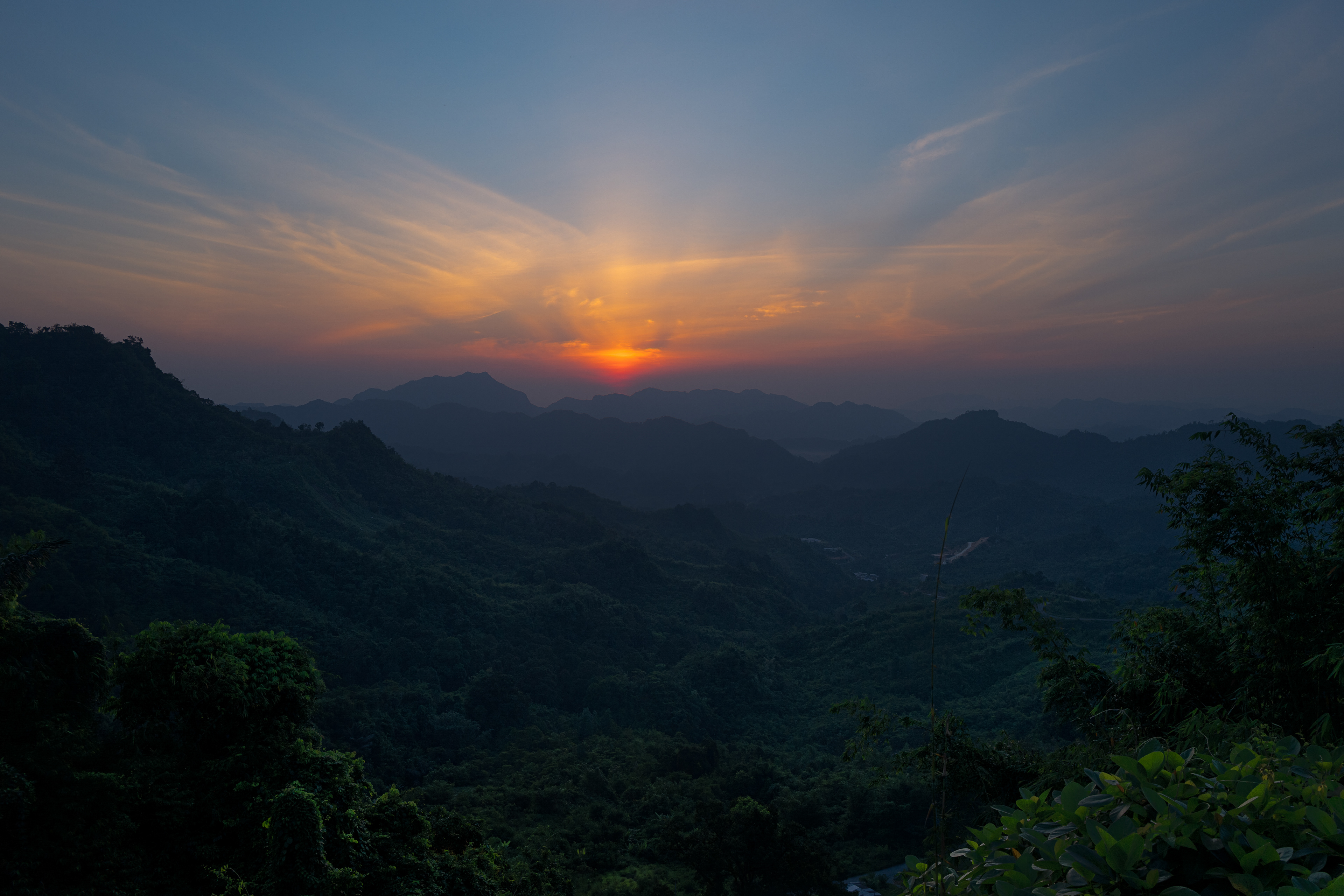 A vivid sunrise breaks above layers of dark, rainforest-covered mountains, casting warm orange and golden light across a hazy horizon. Delicate clouds radiate outward from the rising sun while densely forested valleys remain in deep shadow, creating a dramatic contrast between the awakening sky and the mountainous Borneo landscape.