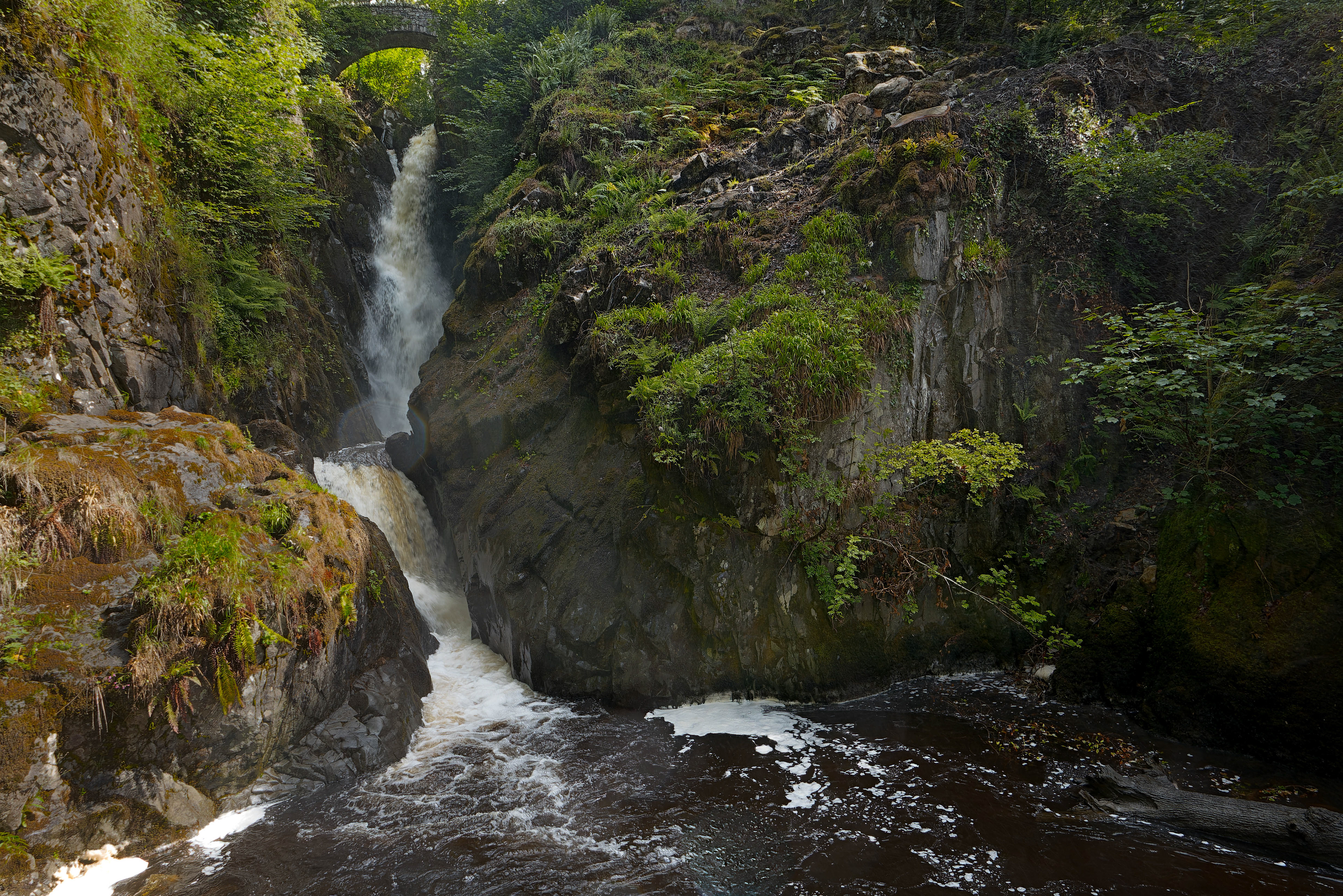 A fine-art landscape photograph of a powerful waterfall plunging through a steep, dark rocky gorge covered in lush green ferns, with a small stone bridge visible at the very top and a churning, tannin-stained pool at the bottom.