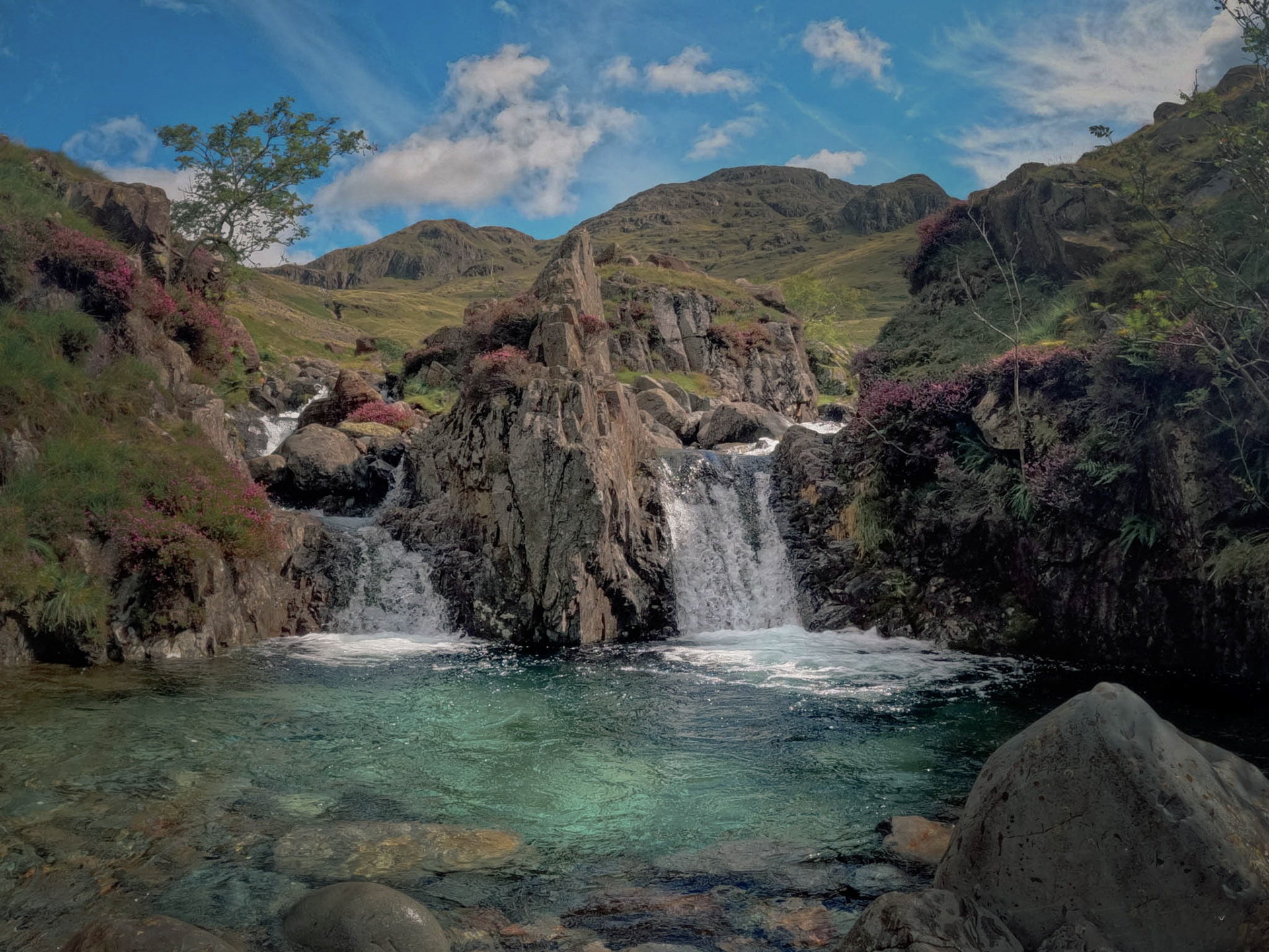 Bright blue sky and cloud frame twin waterfalls spilling into vivid turquoise depths at Wasdale Emerald Pool, ringed by heather-clad rocks and backed by rolling fells.