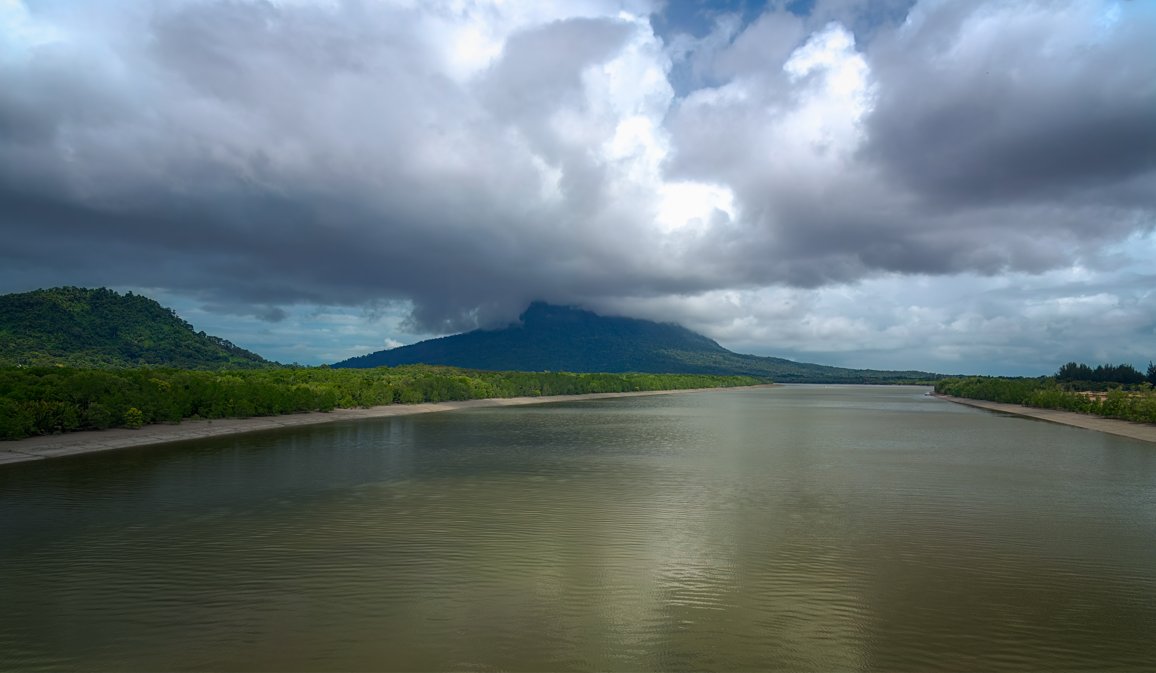 A dramatic fine-art landscape photograph of a wide, calm river leading toward a distant, dark green mountain that is completely enveloped by massive, heavy storm clouds.