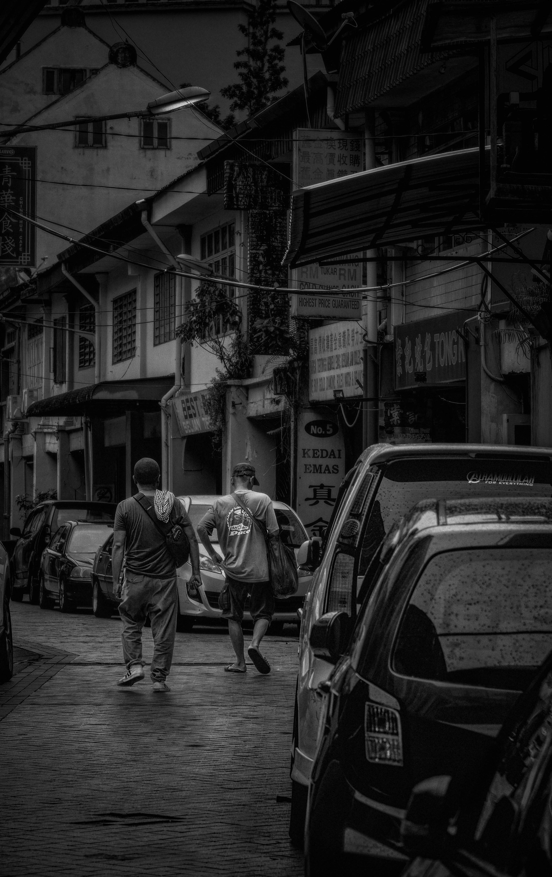 A moody, black-and-white street photograph showing the backs of two men walking down a historic, narrow alleyway lined with parked cars, old shophouses, and vintage storefront signage.