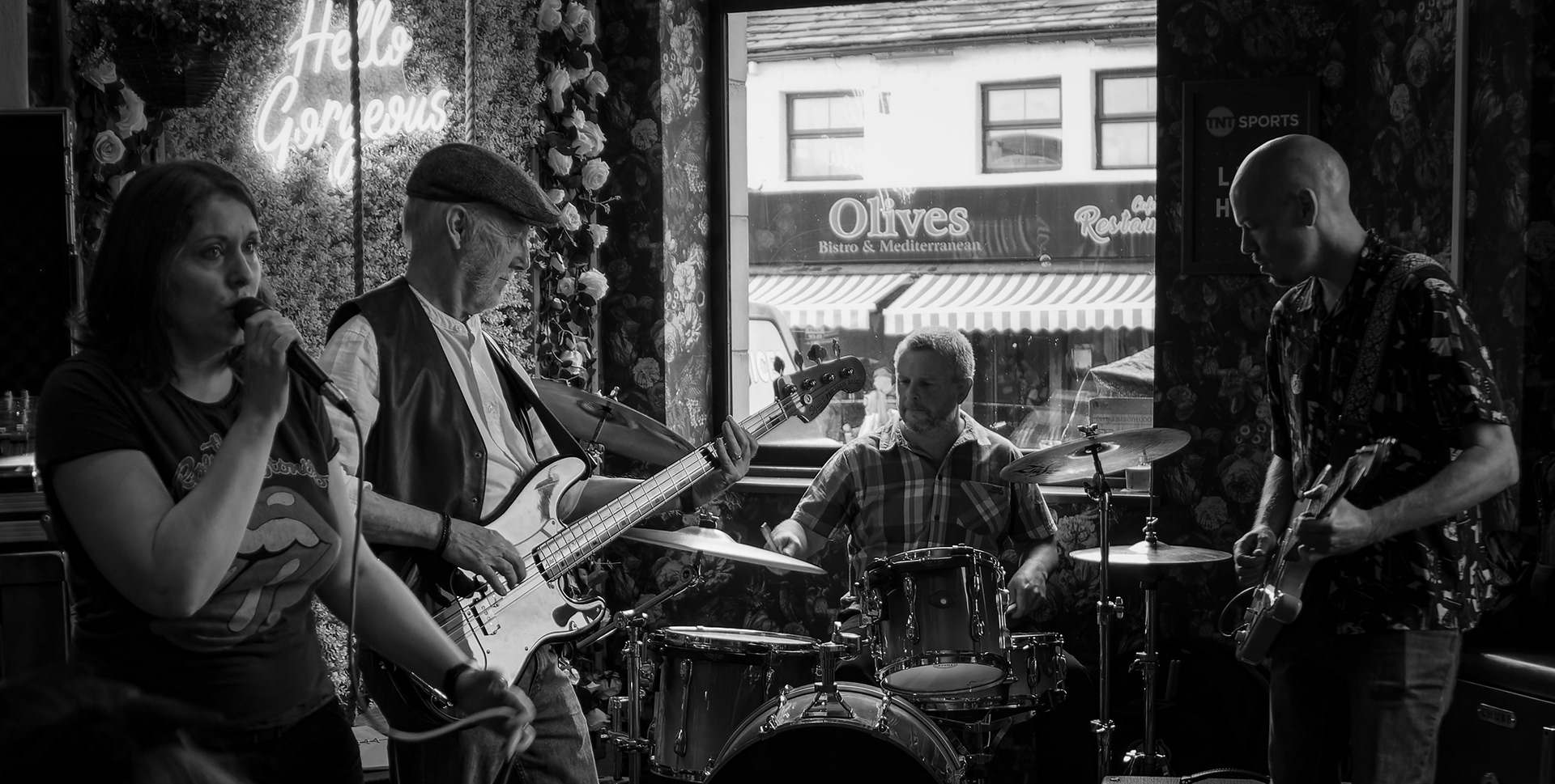 A black and white photograph of a four-piece band performing intimately inside a venue. A female vocalist sings passionately on the left, accompanied by a bassist, drummer, and guitarist, all deeply focused, with a large window behind them revealing a street and a bistro across the way.