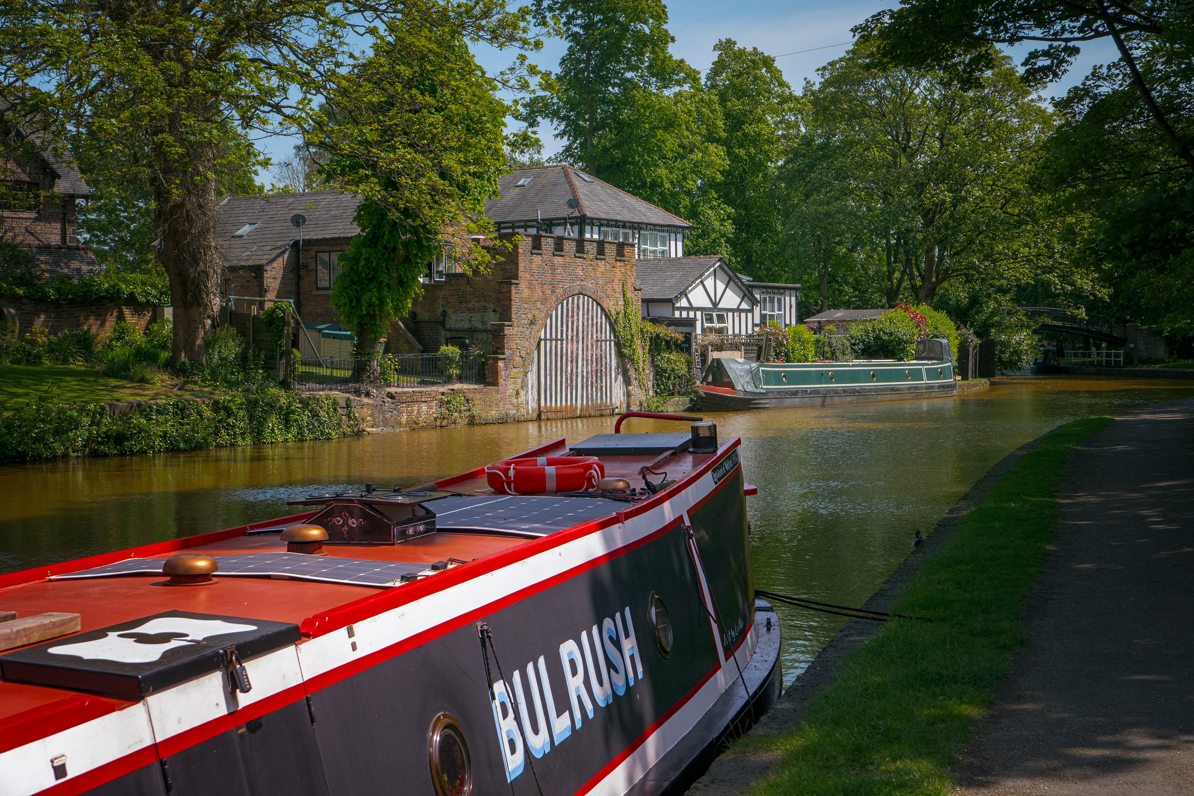 A fine-art landscape photograph featuring a bright red and black narrowboat moored on a calm canal, looking across the water toward a historic brick boathouse, timber-framed buildings, and lush green trees under a bright blue sky.