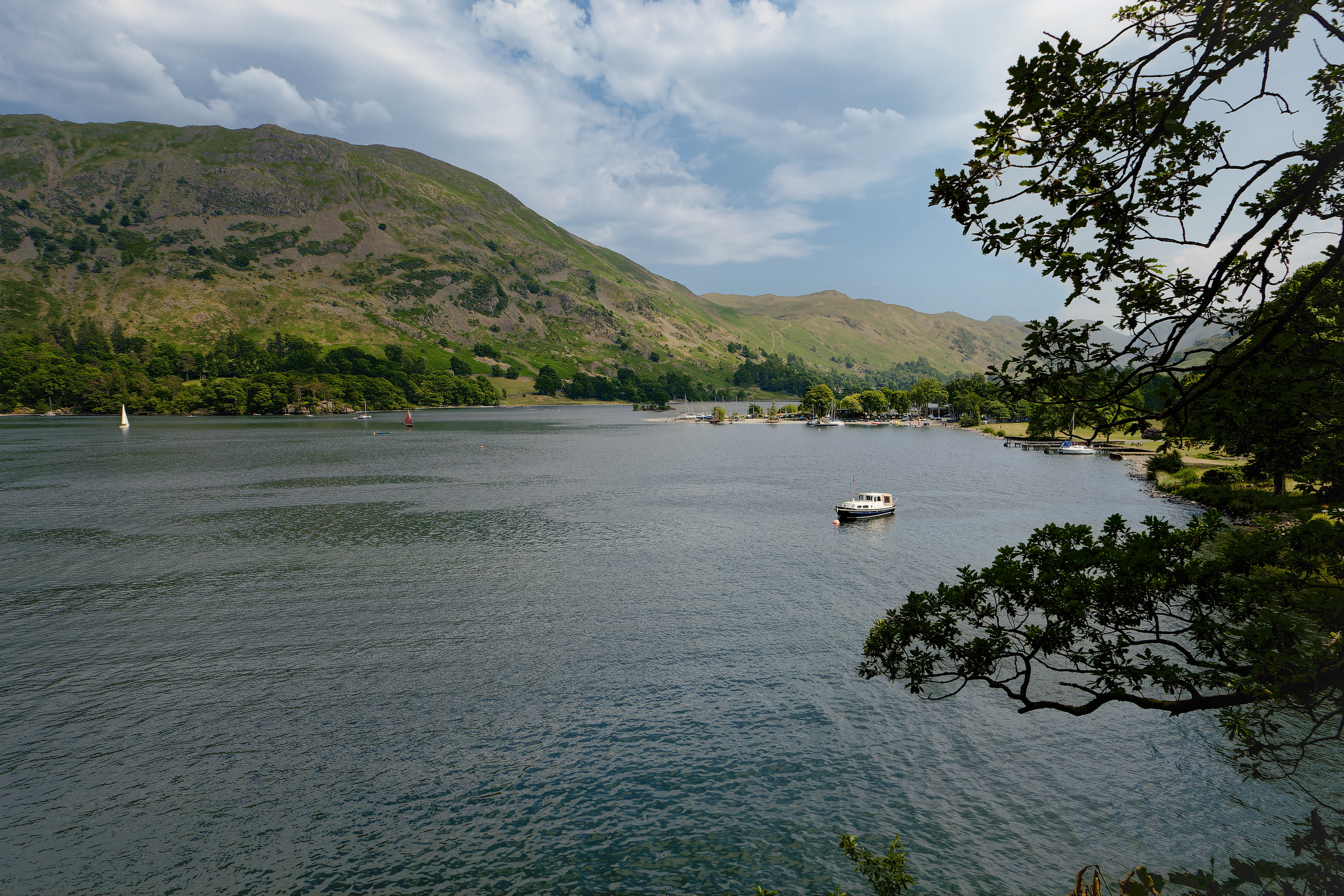 A fine-art landscape photograph showing a vast, rippling lake framed by dark, overhanging tree branches in the foreground, with a single small moored boat in the middle ground and large, rolling green fells rising beneath a dramatic, cloudy sky in the distance.