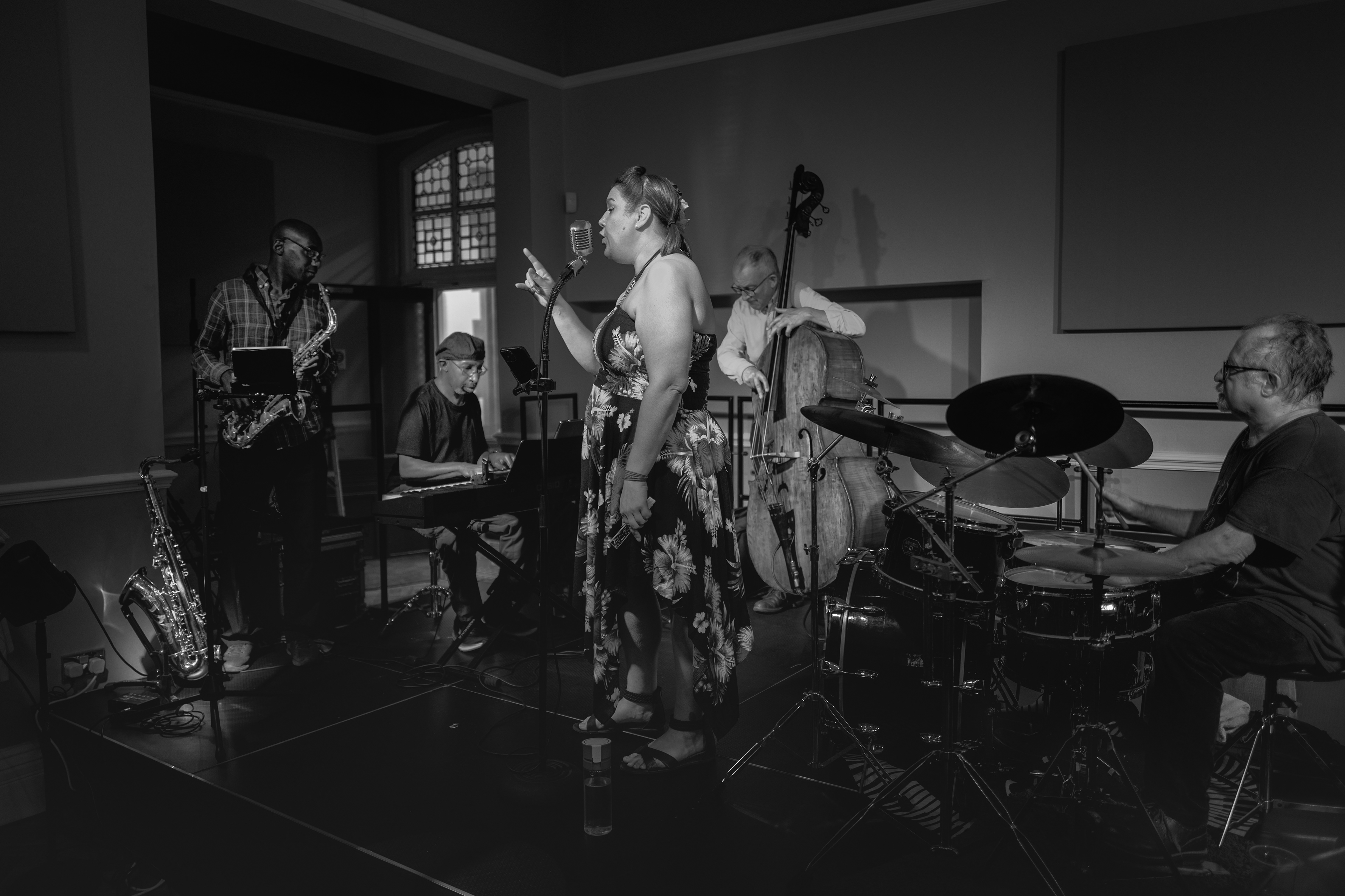 A moody, black and white photograph of a live band performing on a dimly lit stage, featuring a female vocalist in a floral dress passionately singing into a vintage microphone, flanked by a saxophonist, keyboardist, upright bassist, and drummer in deep concentration.