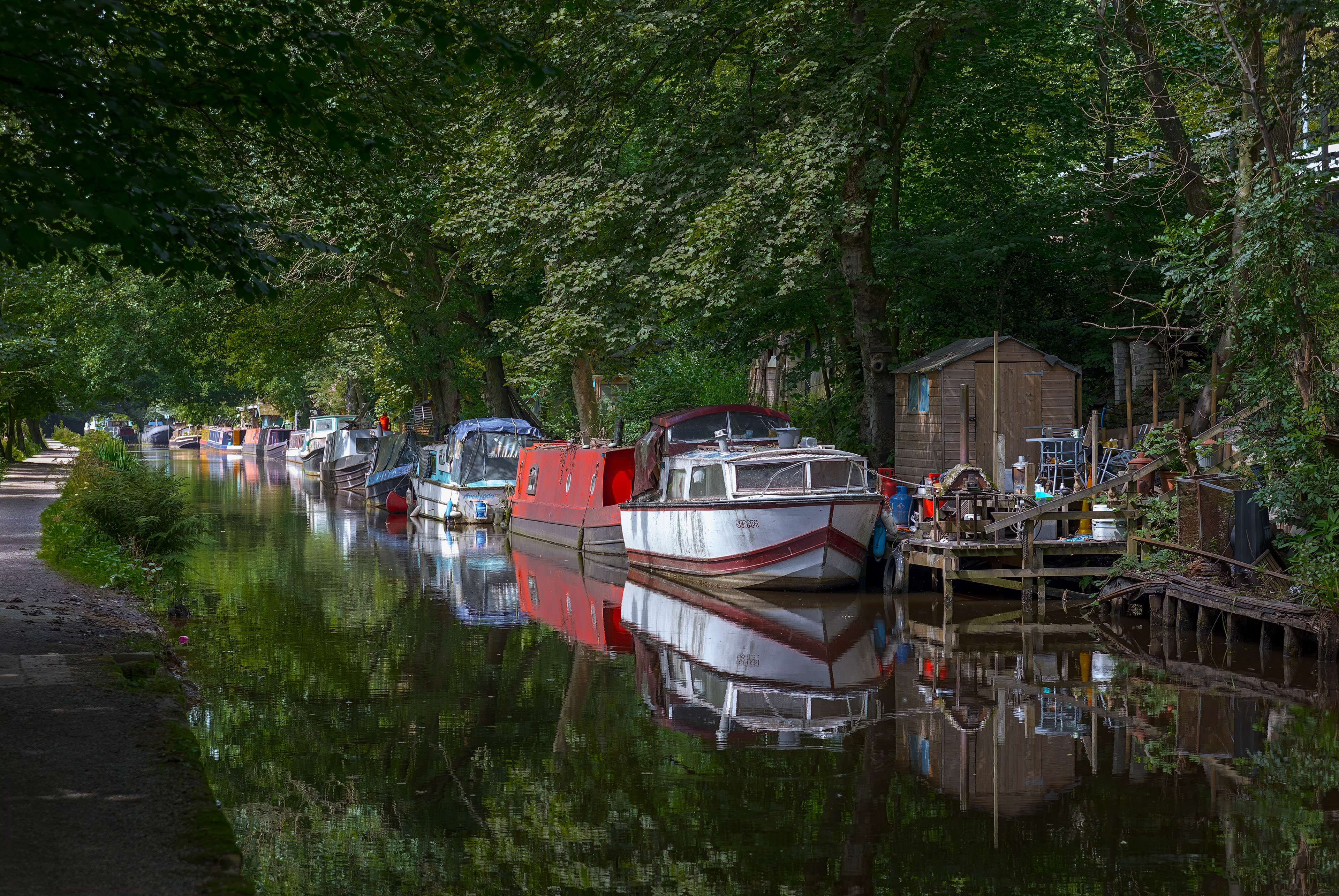 A fine-art photograph depicting aged narrowboats moored along a dark, calm canal, heavily shaded by a canopy of dense green trees whose leaves reflect flawlessly in the mirror-like water below.