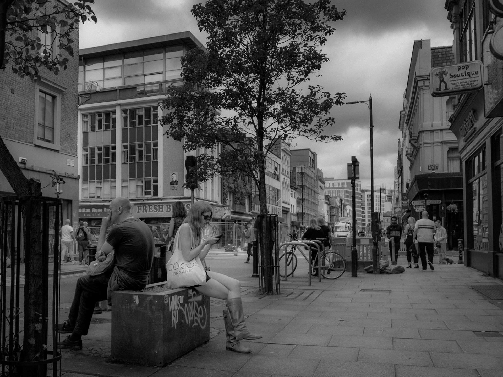 A moody, black and white street photograph depicting a man looking away and a woman looking at her phone, sitting at opposite ends of a graffitied concrete planter on a busy city sidewalk, surrounded by historic buildings, passing pedestrians, and a central tree beneath a dramatic, overcast sky.