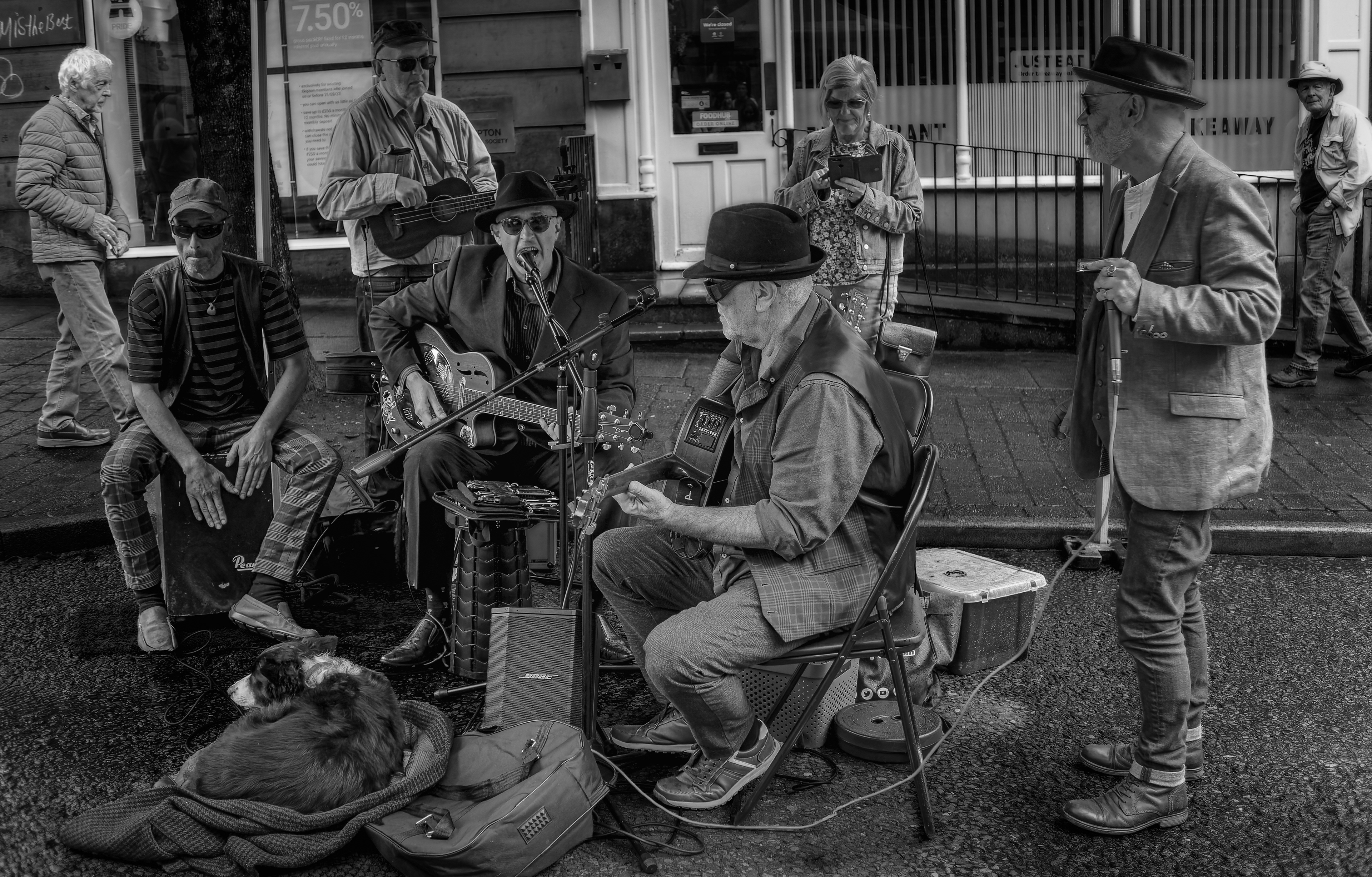 A high-contrast black and white photograph of older street musicians playing a resonator guitar, bass, cajon, and harmonica on a paved sidewalk, with a sleeping dog resting near their instruments and pedestrians in the background.