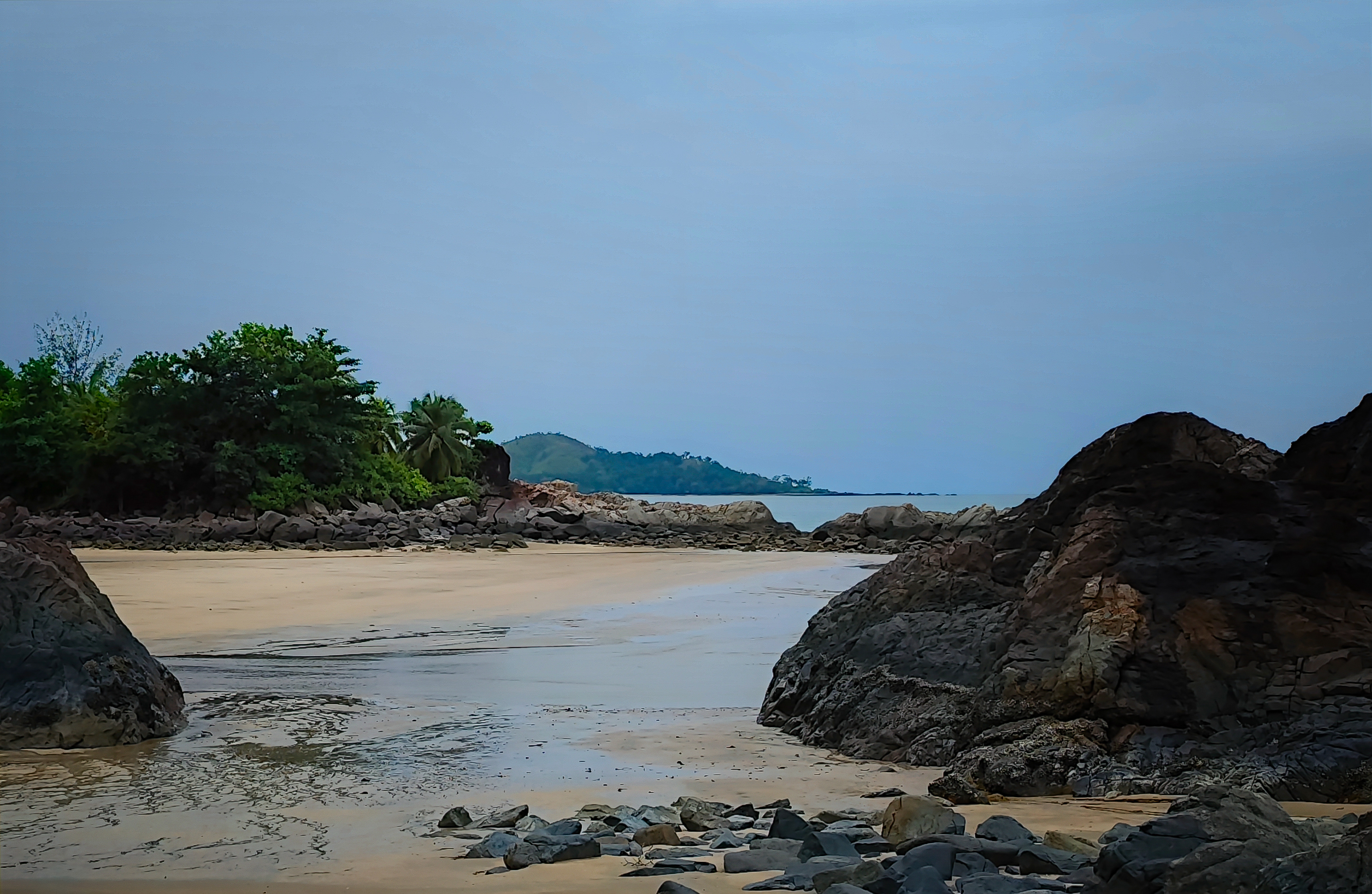 Quiet tropical shoreline framed by dark rocks, with wet sand, calm water and a distant forested headland beneath a muted blue-grey sky.