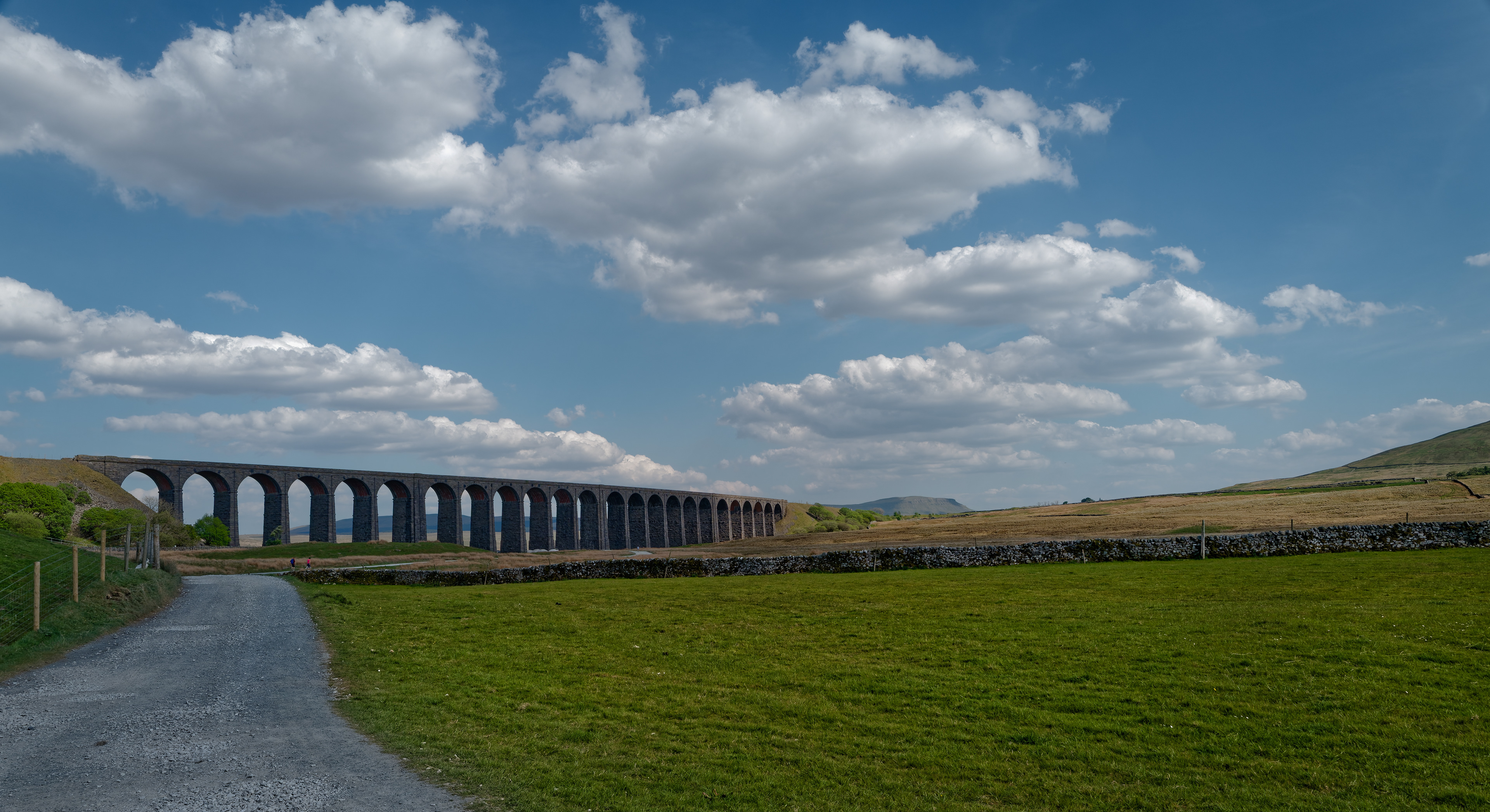 A wide landscape photograph showing a long, multi-arched stone viaduct stretching across a green valley beneath a bright blue sky filled with fluffy white clouds, featuring a gravel path and a dry stone wall in the foreground.