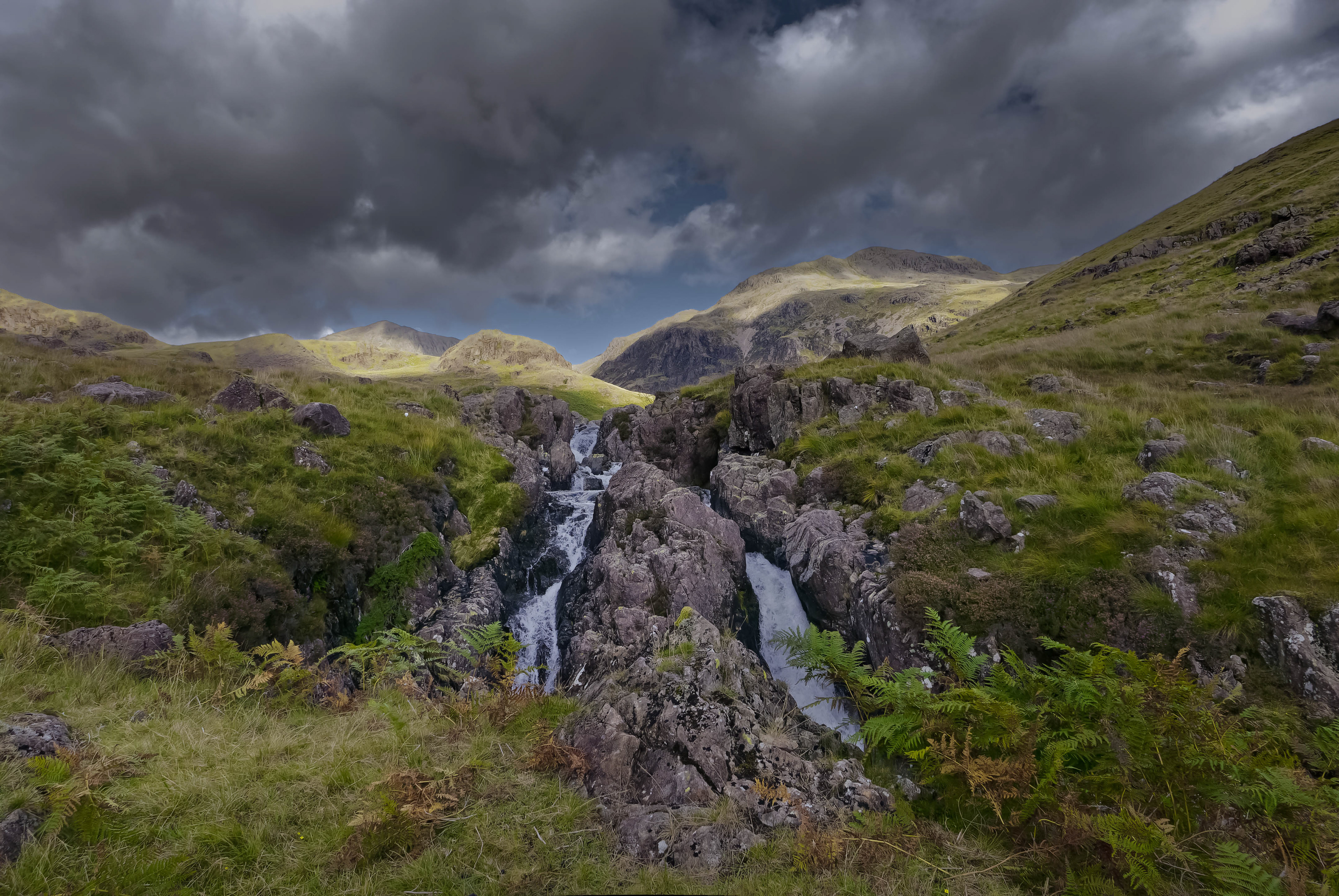 A dramatic landscape photograph featuring a jagged rocky gorge with cascading water in the foreground, leading toward distant sunlit green mountains, all juxtaposed beneath an imposing, dark grey, stormy sky.