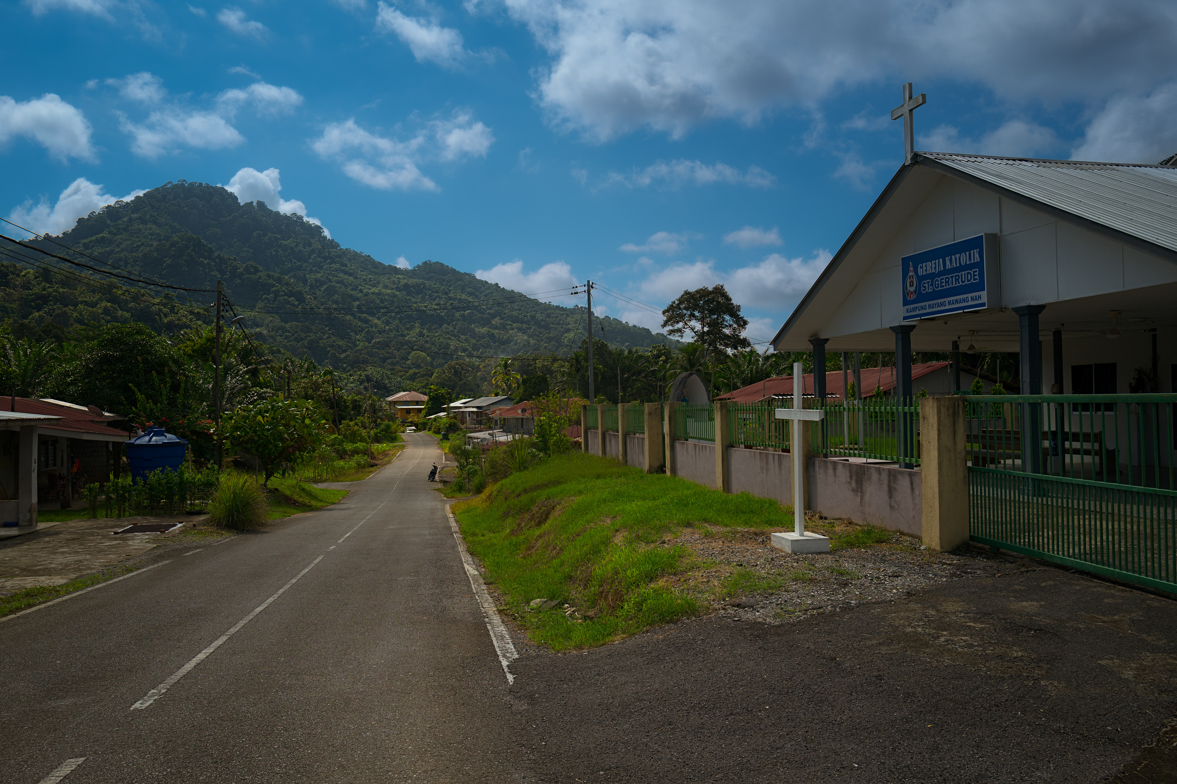 A quiet paved country road runs alongside a small white church with a rooftop cross and a freestanding white cross, leading the eye toward a large, lush green mountain under a vibrant blue sky with scattered clouds.