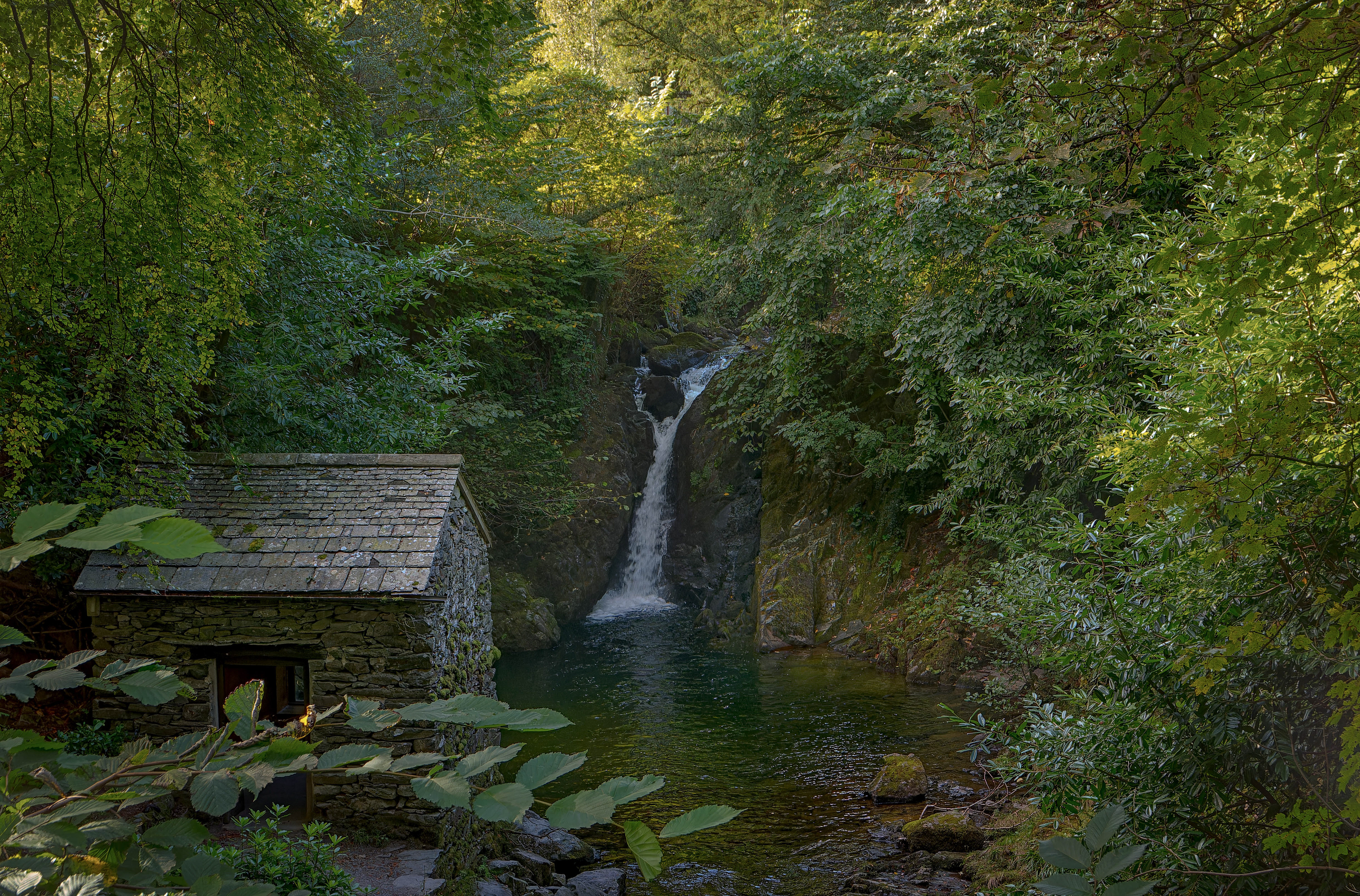 A fine-art landscape photograph of a small waterfall tumbling into a dark, serene woodland pool, bordered by dense, lush green foliage and a small, historic stone building with a slate roof on the left bank.