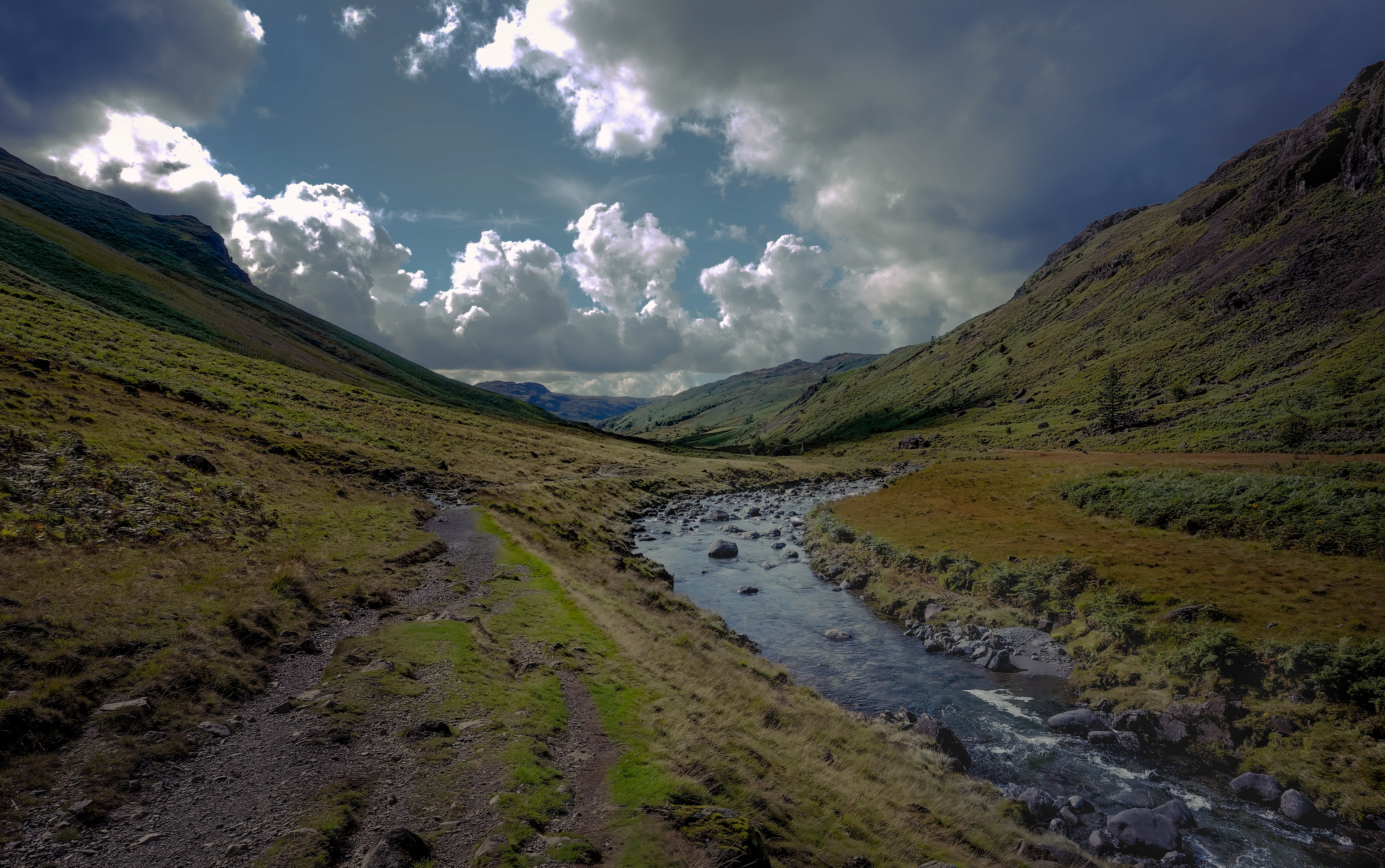 A dramatic, moody landscape photograph of a deep, rugged valley under a stormy sky, featuring a winding stream and a rough path flanked by steep, shadowed mountains.