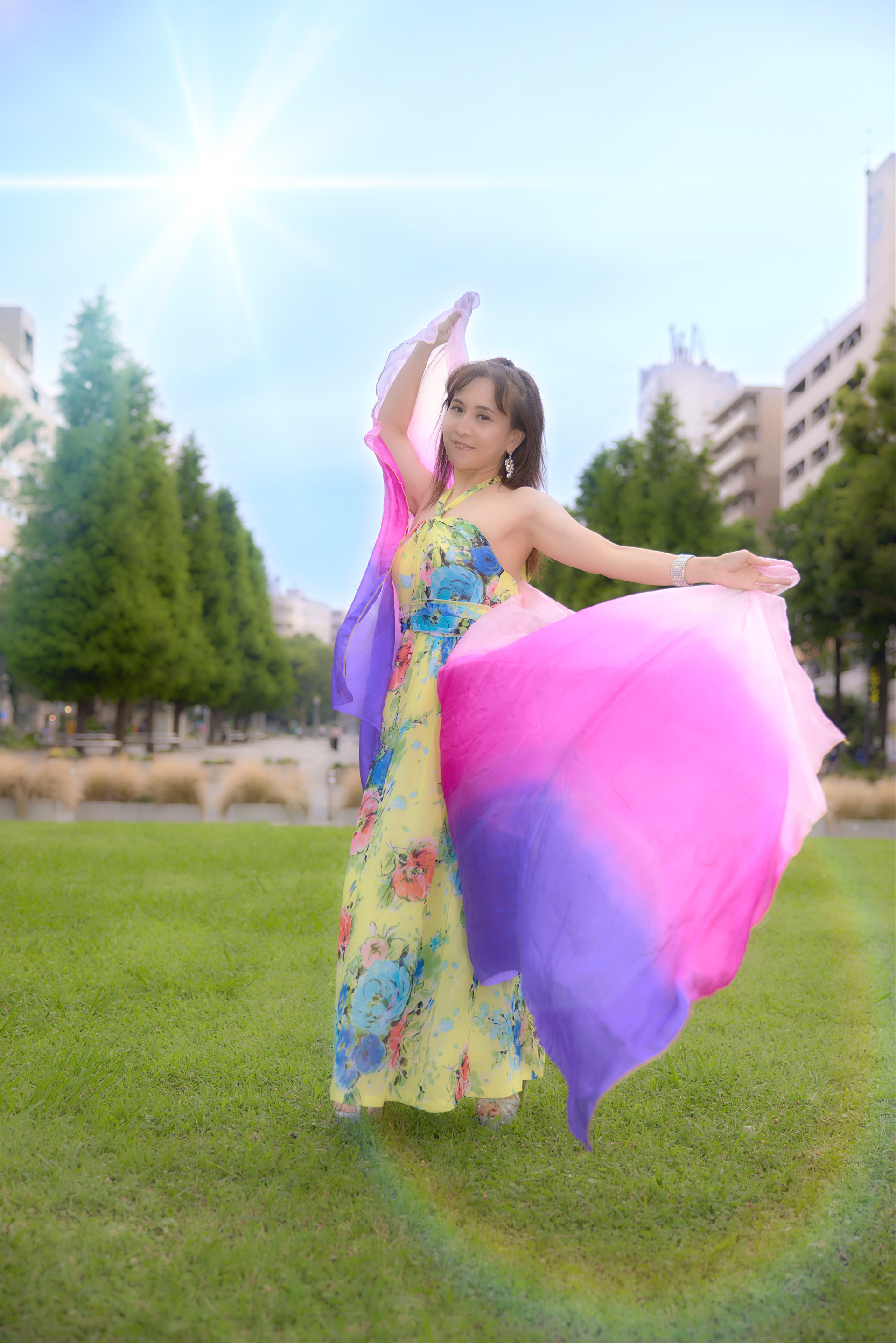 (工夫)日中シンクロ、露出調整、レタッチ、公園の緑の芝生の上で黄色いワンピースとカラフルなストールを羽織っためぐみさんの写真です。曇りがちだったのですが、青空と太陽、そしてフレアを再現させました／(カメラ)CANON EOS R5／(レンズ)CANON RF24-70mm F2.8 L IS USM／(撮影日)2022年7月18日／(撮影場所)関内大通り公園／(モデル)三尾めぐみ