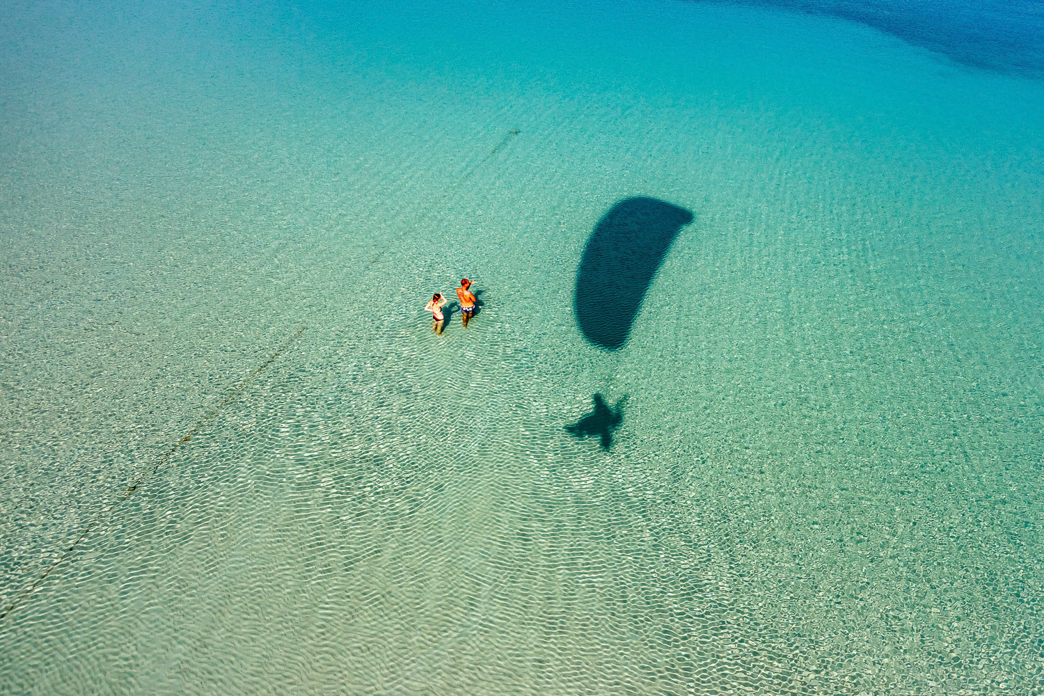"Cesme" - I was in Cesme on the Aegean Sea, a very popular summer town, heading the jury for the national underwater photo contest that year. I drove there with my two paramotors in the back of the car for maybe getting a chance to fly since I had never flown there. I knew there are crazy winds there due to Cesme being on a peninsula with bays, inlets and hills. Being October, air was a little cooler and less turbulent. I found myself a piece of clearance inland to take off. I flew over this couple standing knee deep in crystal clear water on white sandy bottom. My shadow being in the image makes it also a self portrait.