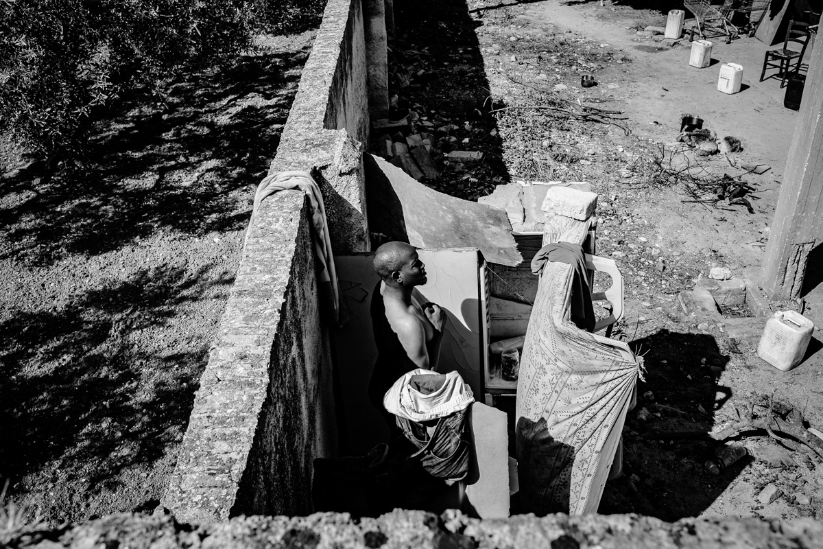 Abdulie, taking bath in front of abounded house, his shelter, in the middle of nowhere. He was living in the informal camp for around 3 years before police came to force all people to leave. Together with his several friends, he moved to this house, there is no electricity, no water, toilet and bathroom. He is in very deep depression, for many months he have no work, also for more than 4 years he doesn't seen his family, wife and 3 children. Also every day, several times, his mum and wife call him online to ask about money for food for all his family.