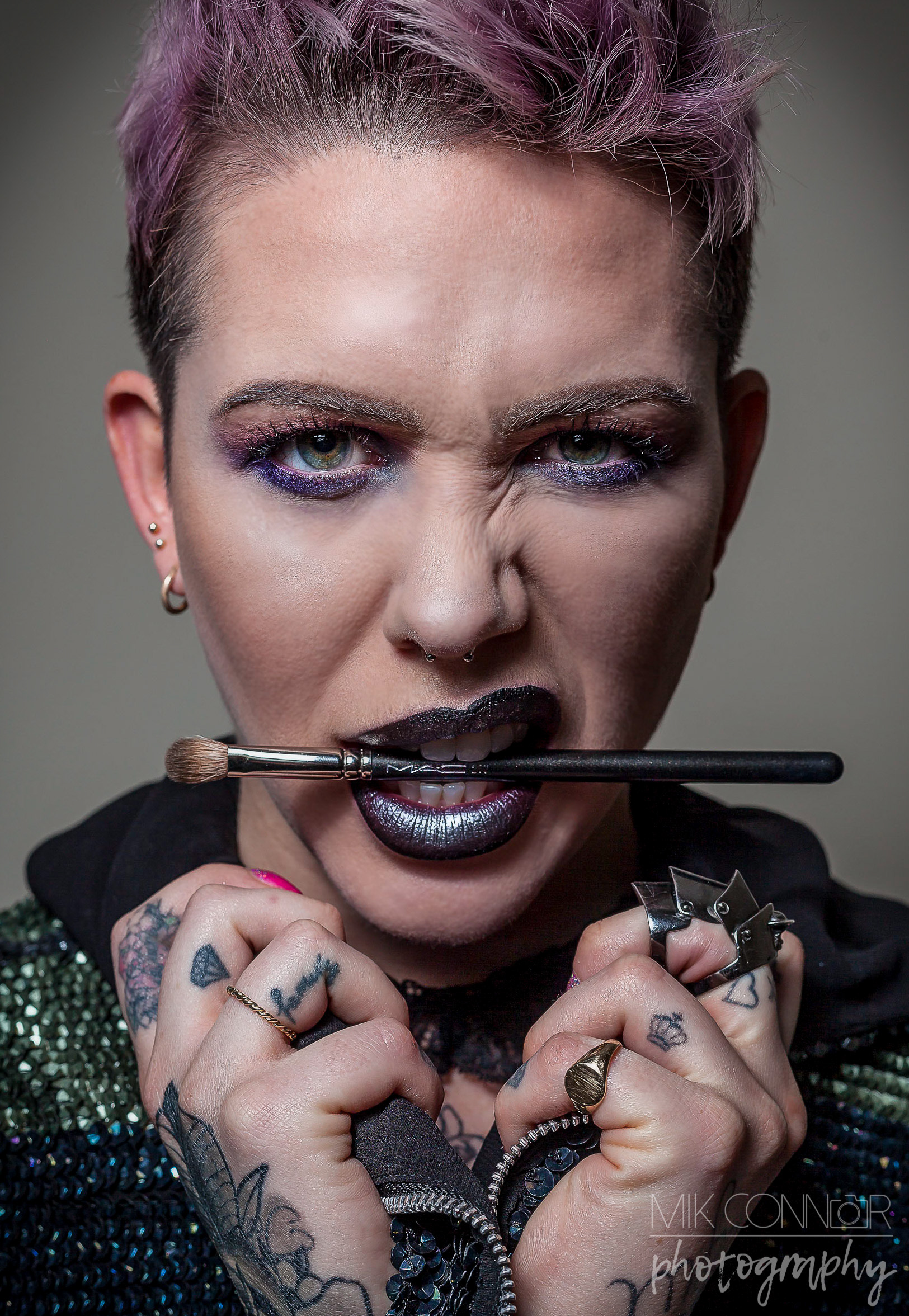 Portrait of an alternative type female model posing with the hands gripping the collar of her hoodie to show of her tattoos, snarling with a make up brush between her teeth