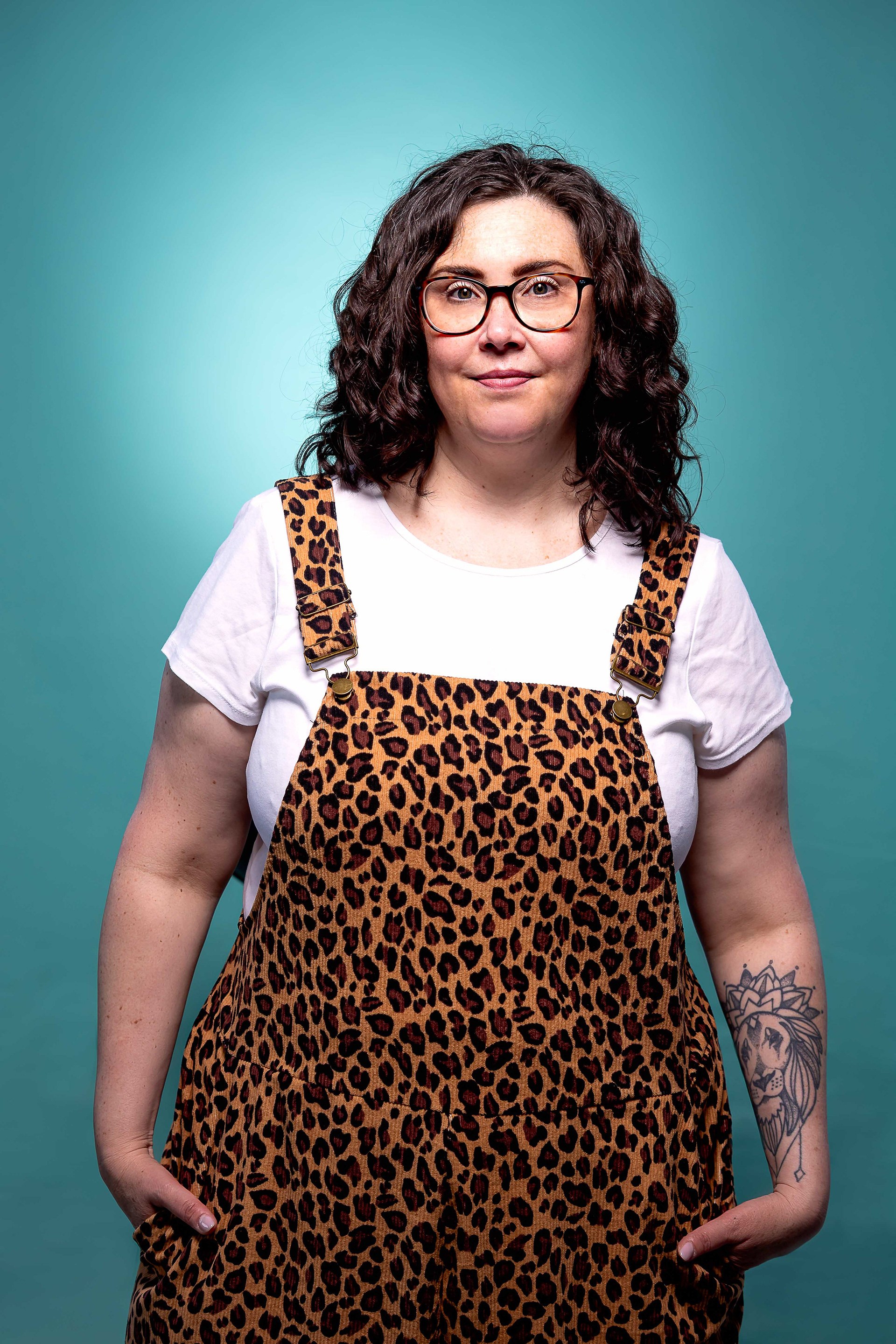 Portrait of a dark haired woman wearing a white t-shirt under leopard print dungarees in front of a light blue background