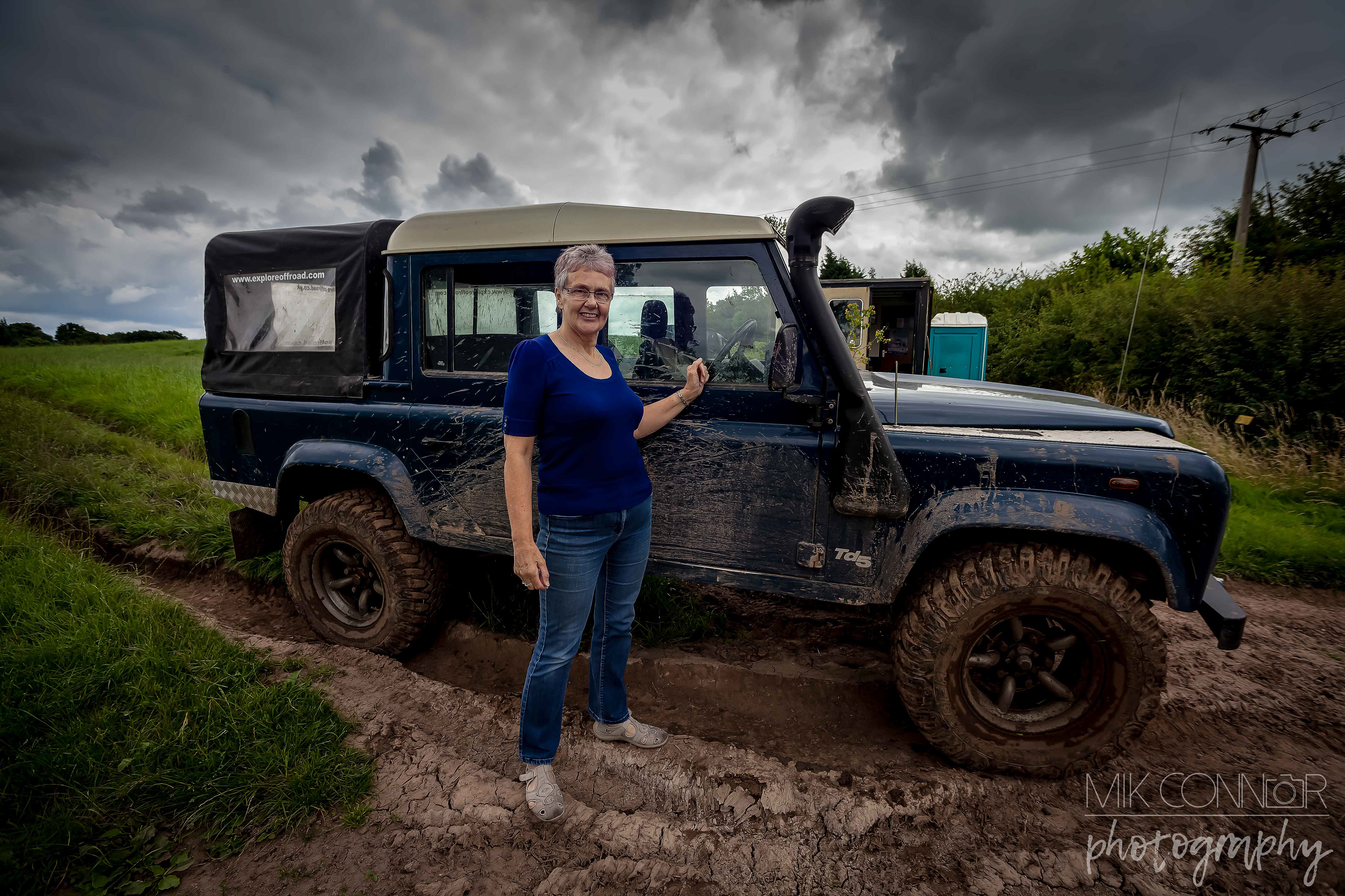 Wide angled portrait of a woman stood next to a land rover in a muddy field while rain clouds gather above