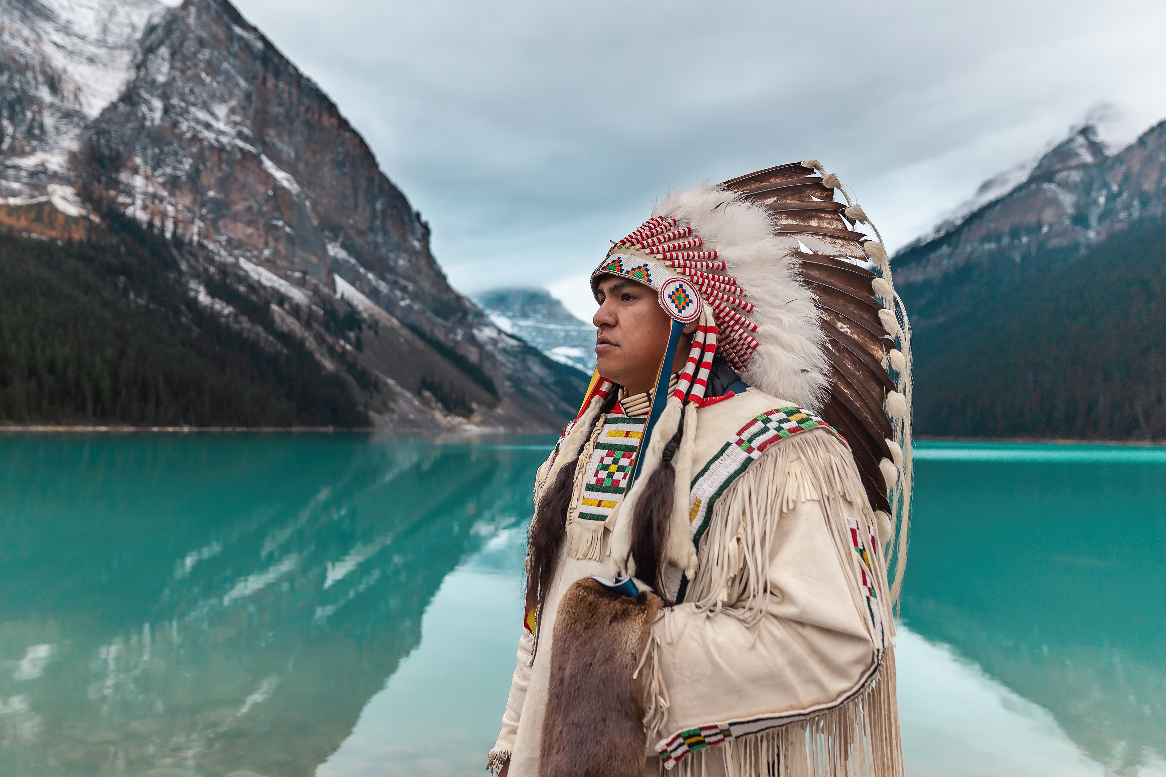 Portrait of a first nations canadian posed looking into the distance wearing full traditional head dress infront of mountains and a lake 