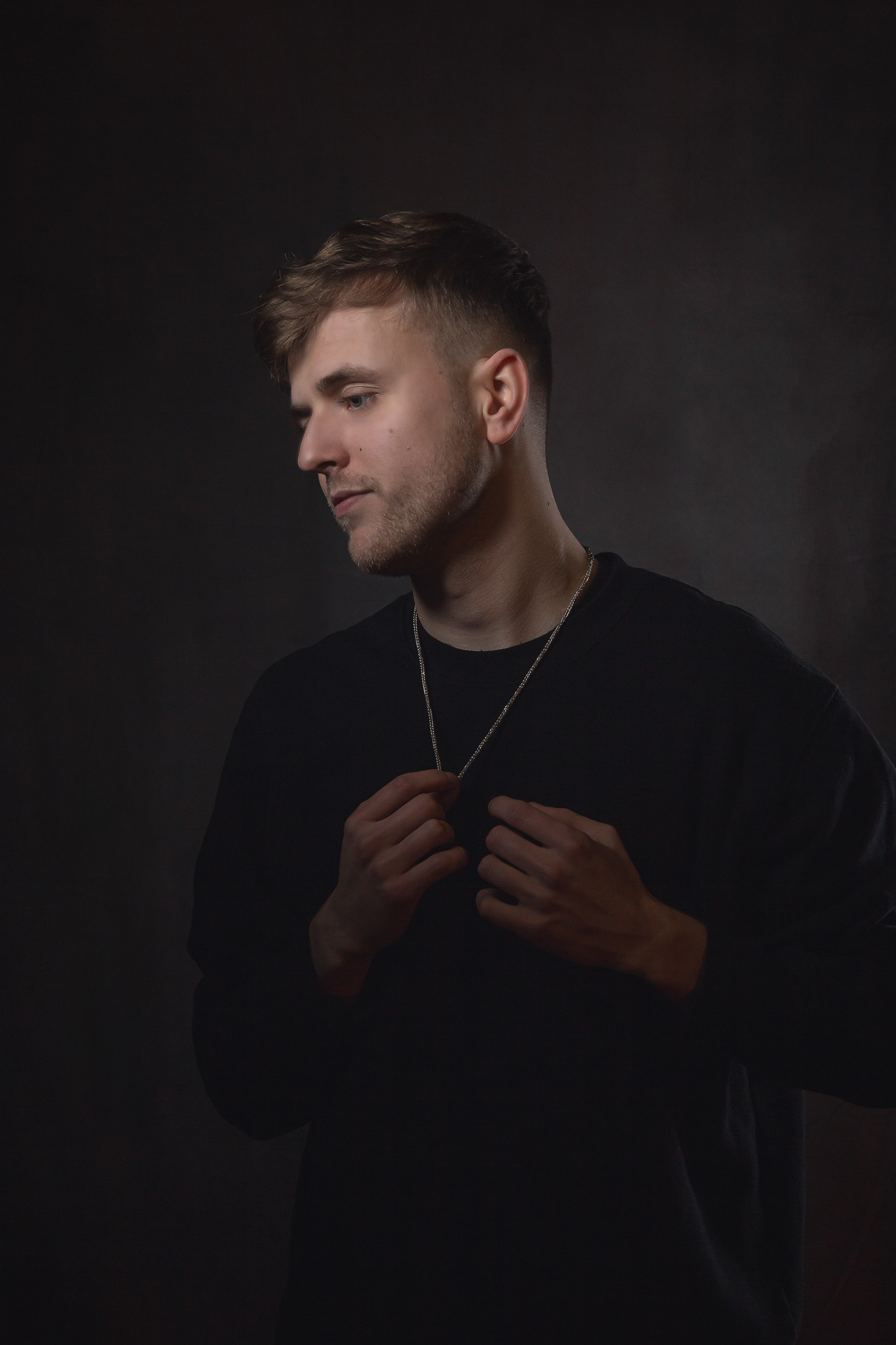 Dark and moody portait of a man looking away from the camera while he rearranges his necklace
