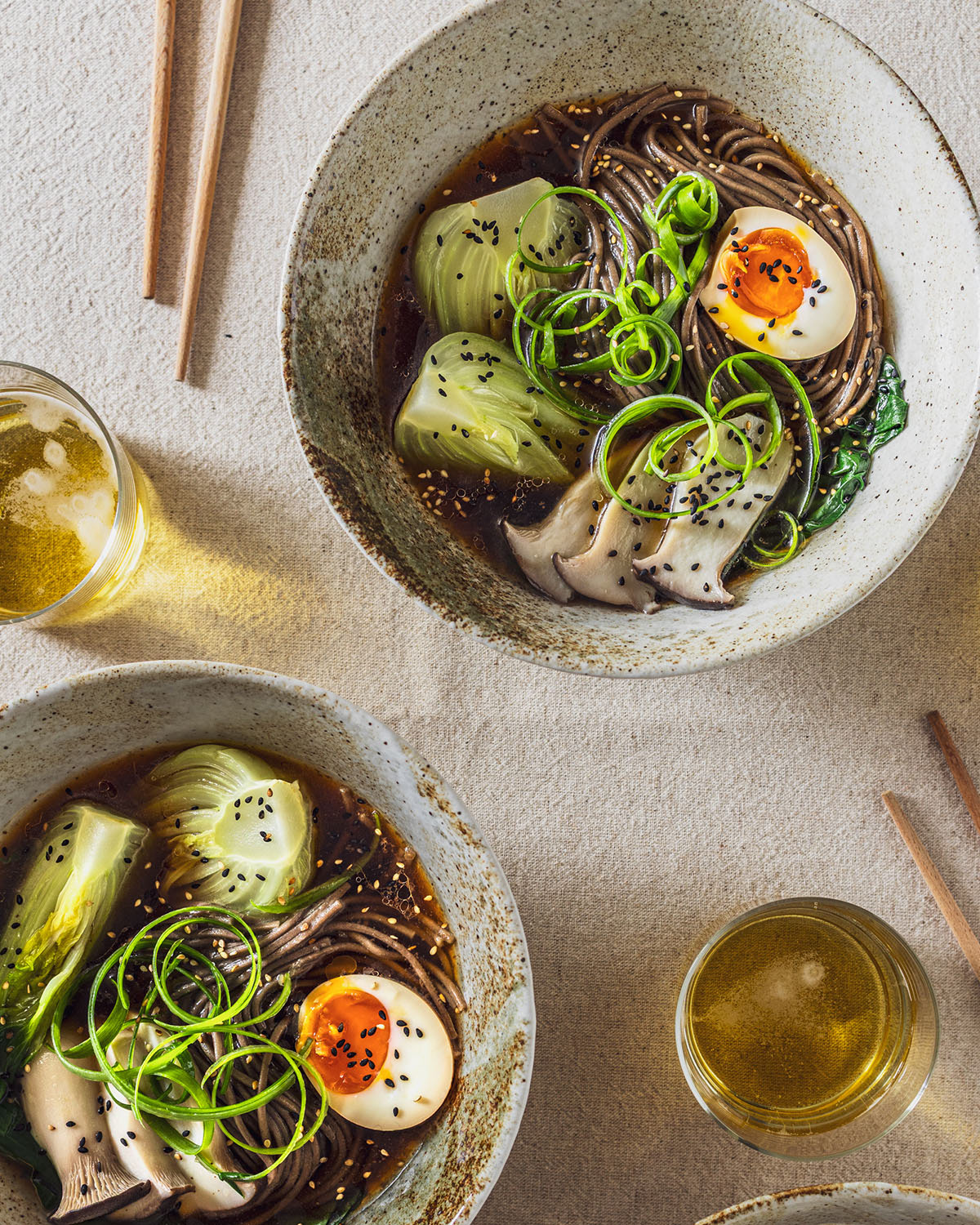 View from top of two bowls or ramen soup with soba noodles, pak choi, mushrooms and soy sauce egg halves. On the side chopsticks and glasses of beer.