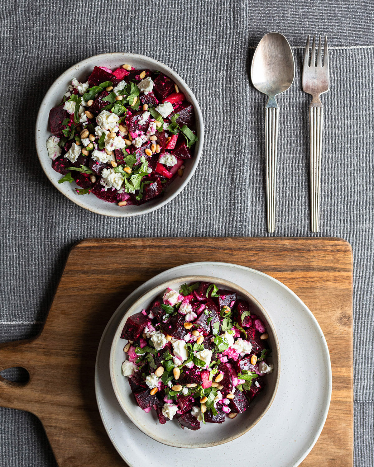 Set table with wooden board and white tableware, beetroot salad with feta, pine nuts and fresh coriander, silver cutlery on the side, grey tablecloth.