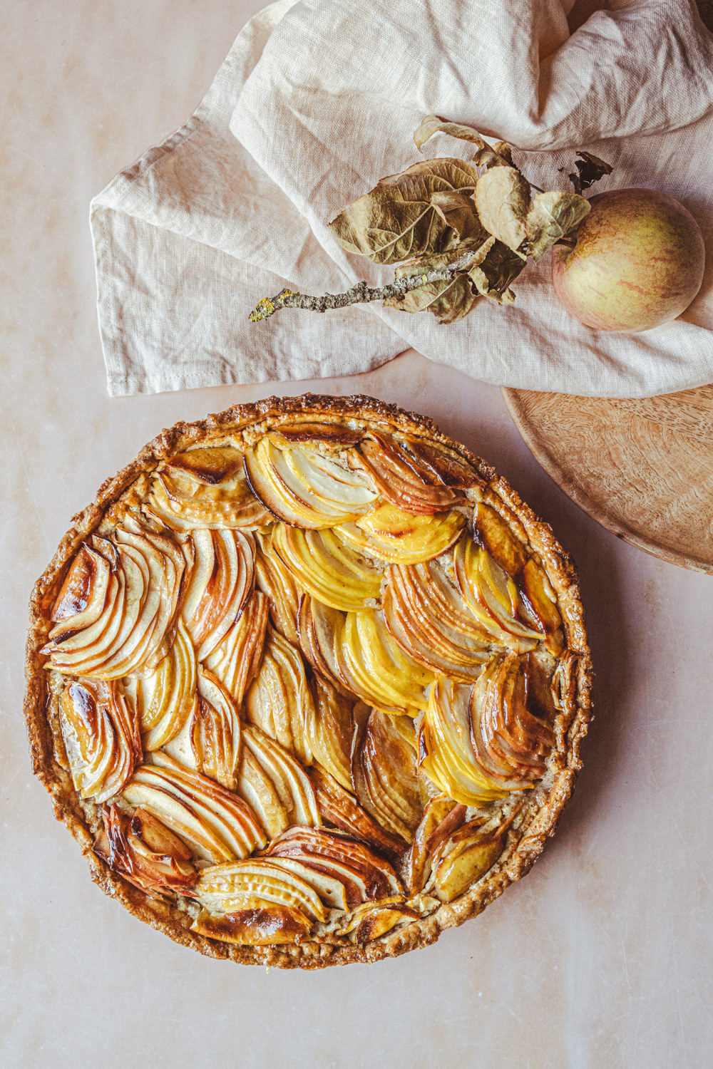 Top view of apple tart. Apples are sliced and arranged in pattern. Piece of cloth and apple on twig as decoration next to it.