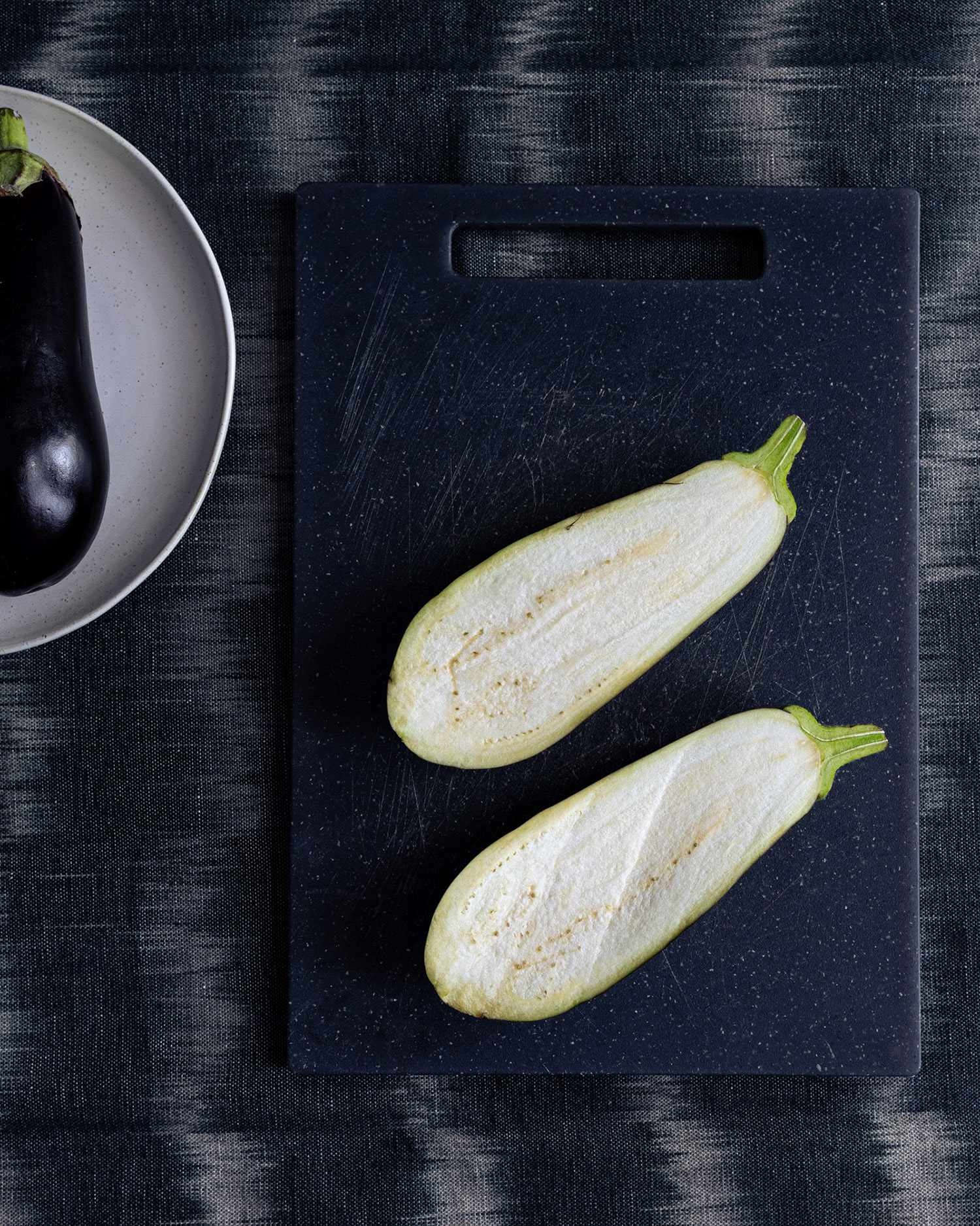 Two eggplant halves on a dark grey cutting board on dark table cloth.