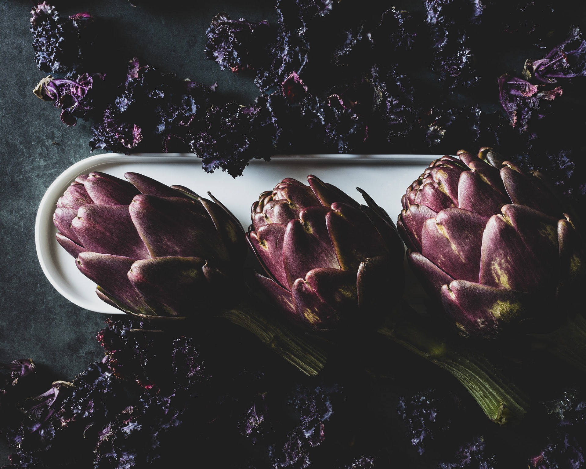 Three purple artichokes on an oval shape white porcelain plate surrounded by purple brussel sprouts.