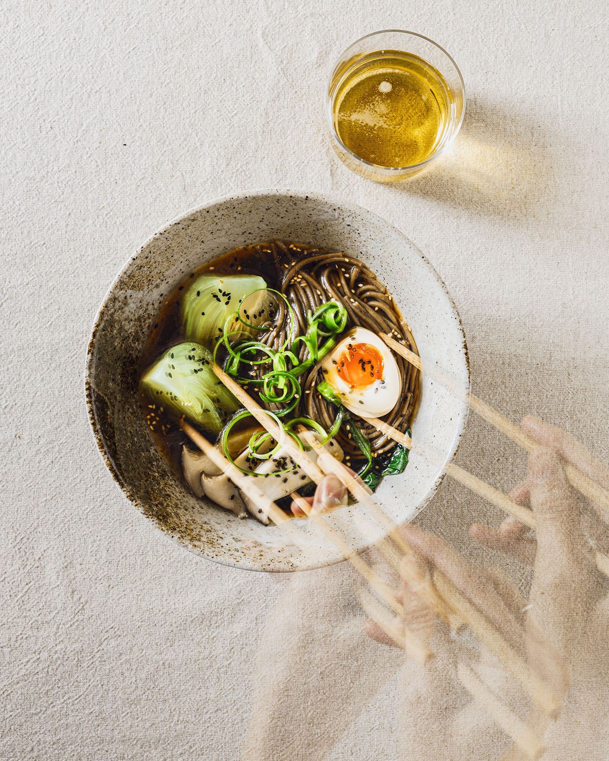 A bowl of ramen noodles with pak choi, soy sauce egg half, and mushrooms on a table. A hand with chopsticks is captured in multiple movements. A glass of beer is on the side.