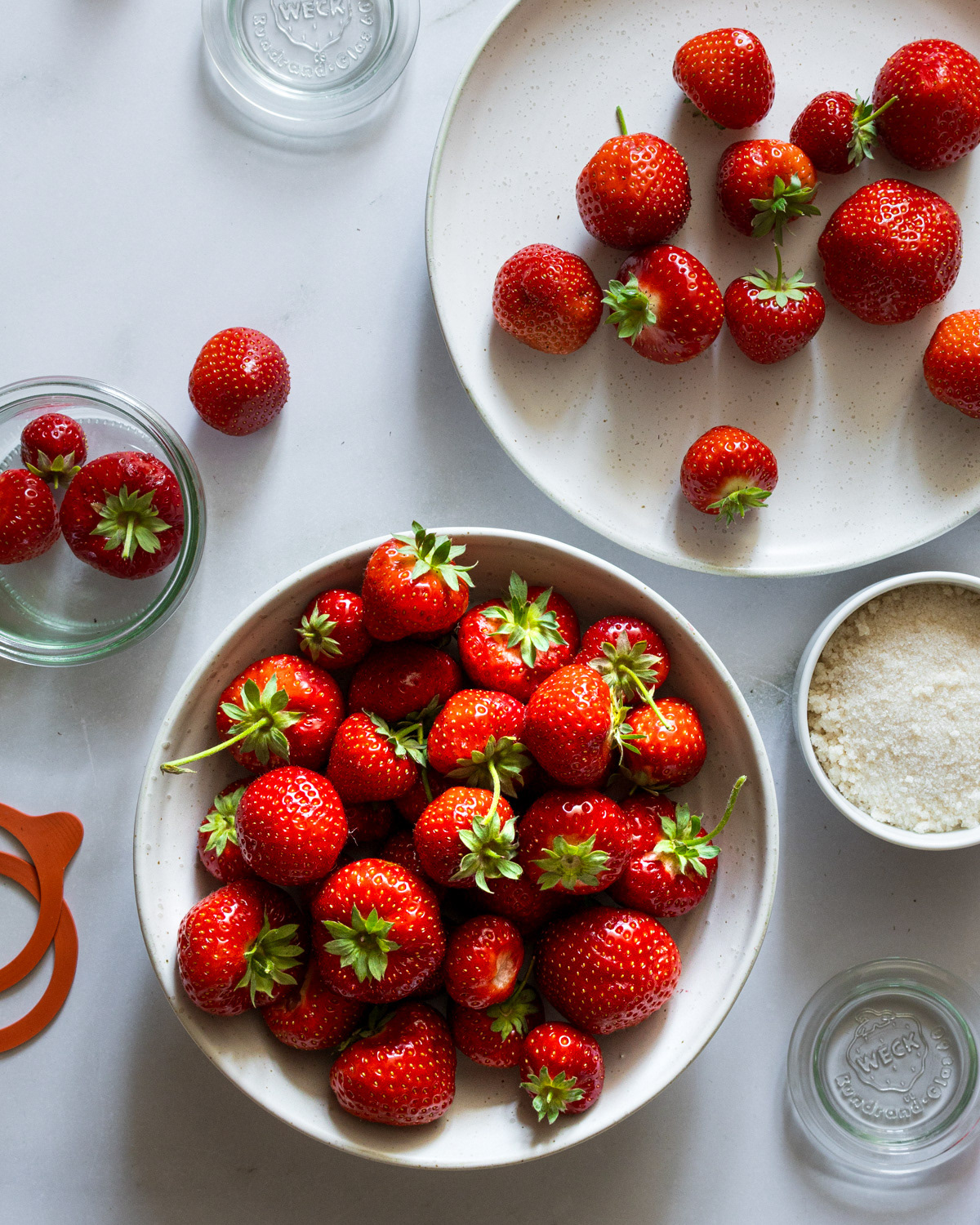 Ripe red delicious strawberries in bowls next to jam making utensils.