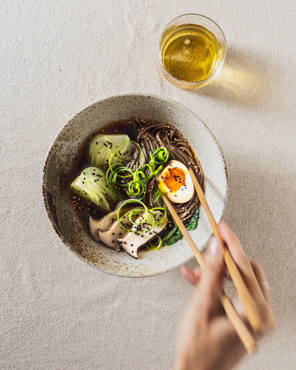 Ramen bowl with soba noodles, pack choi, mushrooms and soy sauce egg halves. A hand in movment is picking up the egg with chopsticks. A glass of beer is on the side.