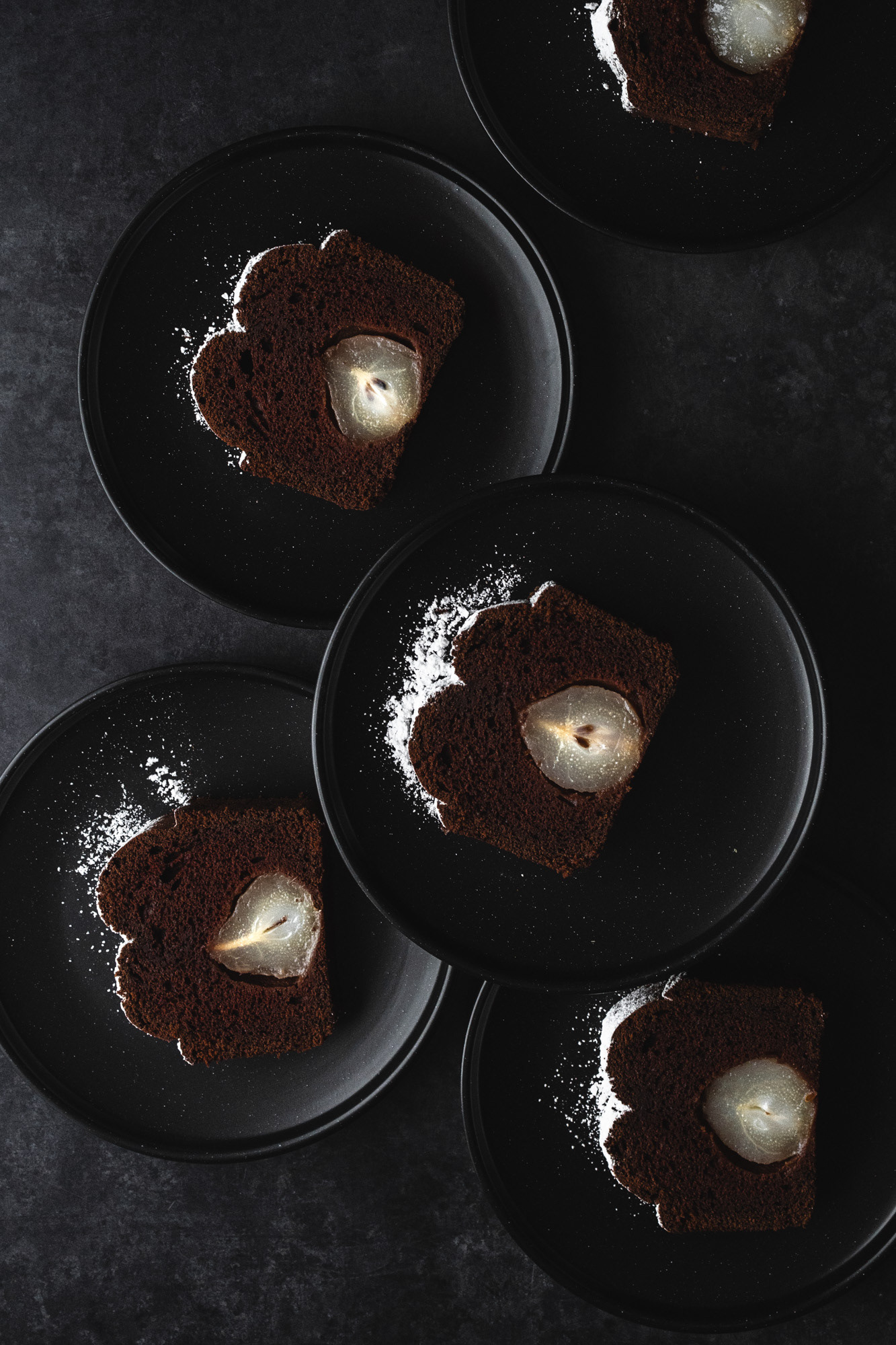 Slices of chocolate pear cake with icing sugar on black plates and dark background creating a pattern. 
