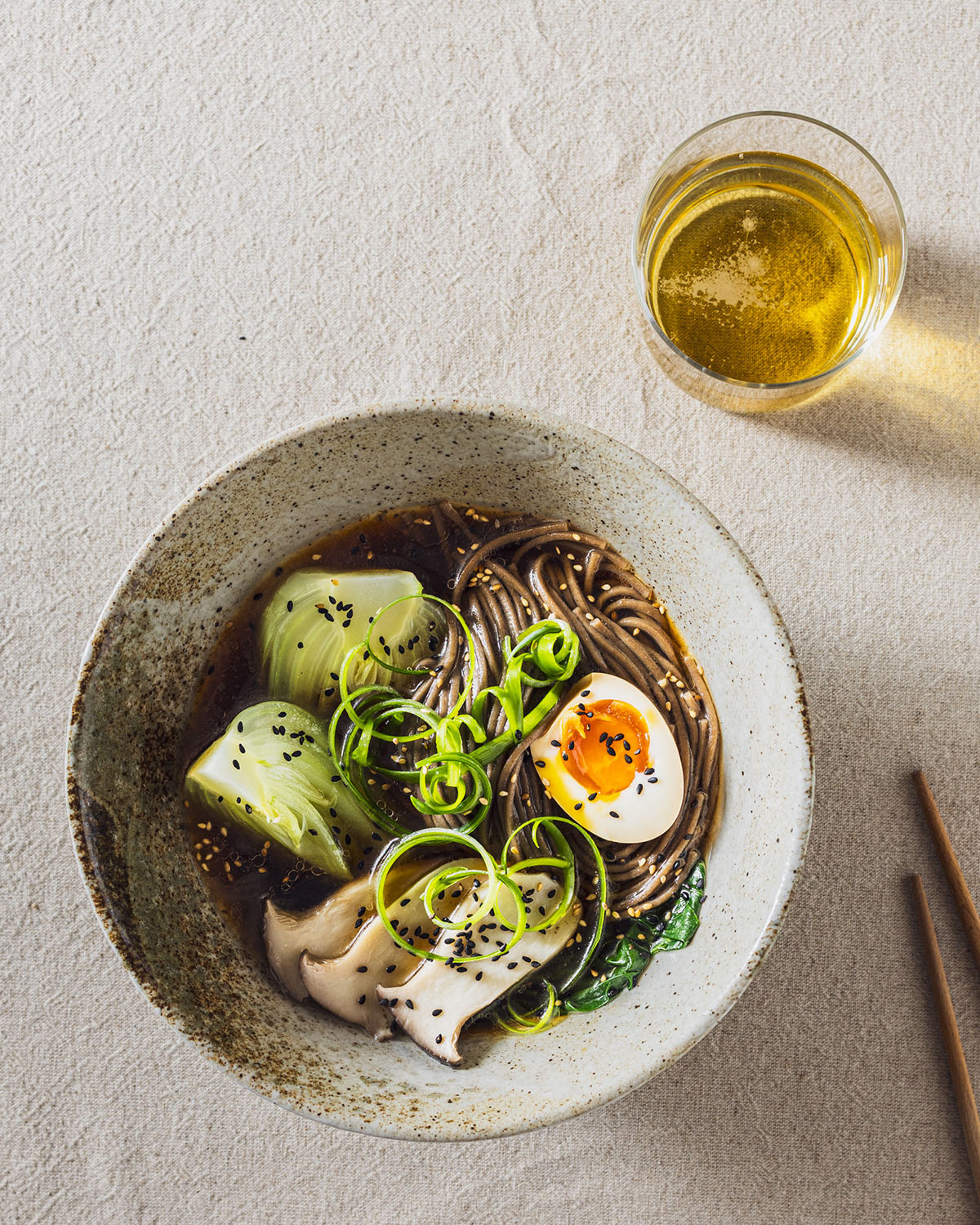 A ramen bowl with soba noodles, pak choi, mushrooms and soy sauce egg halves. Chopsticks and a glass of beer are next to the bowl.