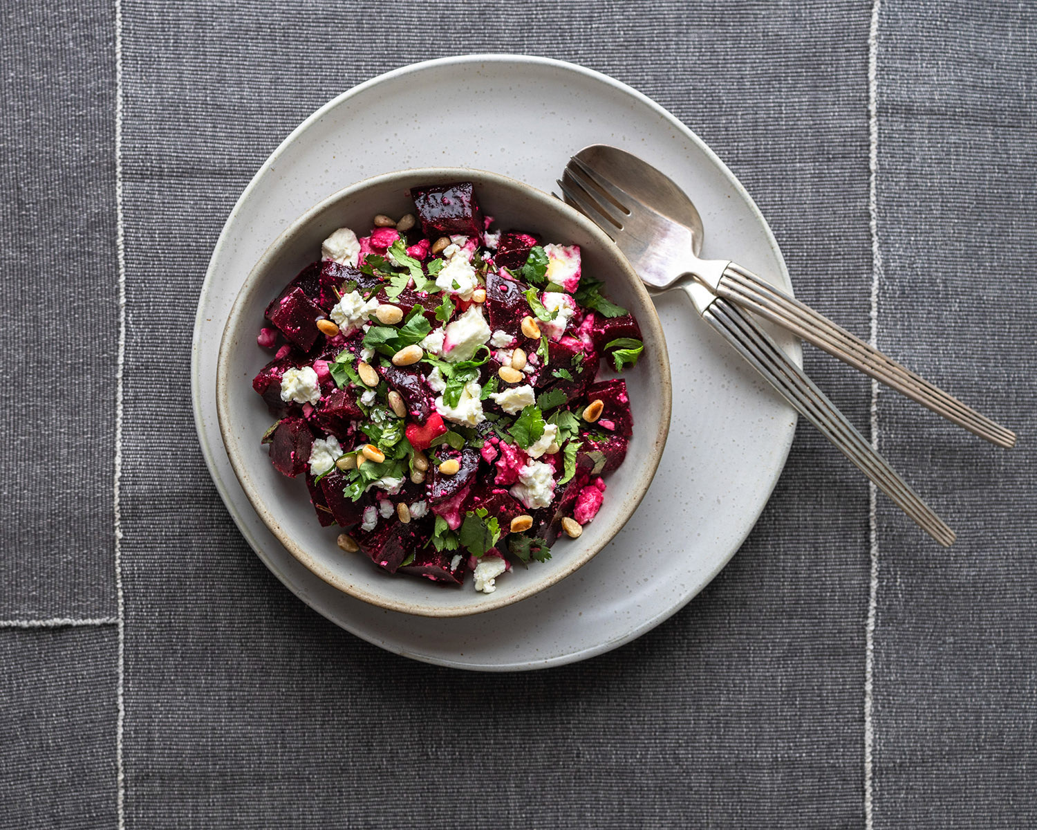 Beetroot salad with feta, pine nuts and fresh coriander in a bowl with cutlery on grey tablecloth.
