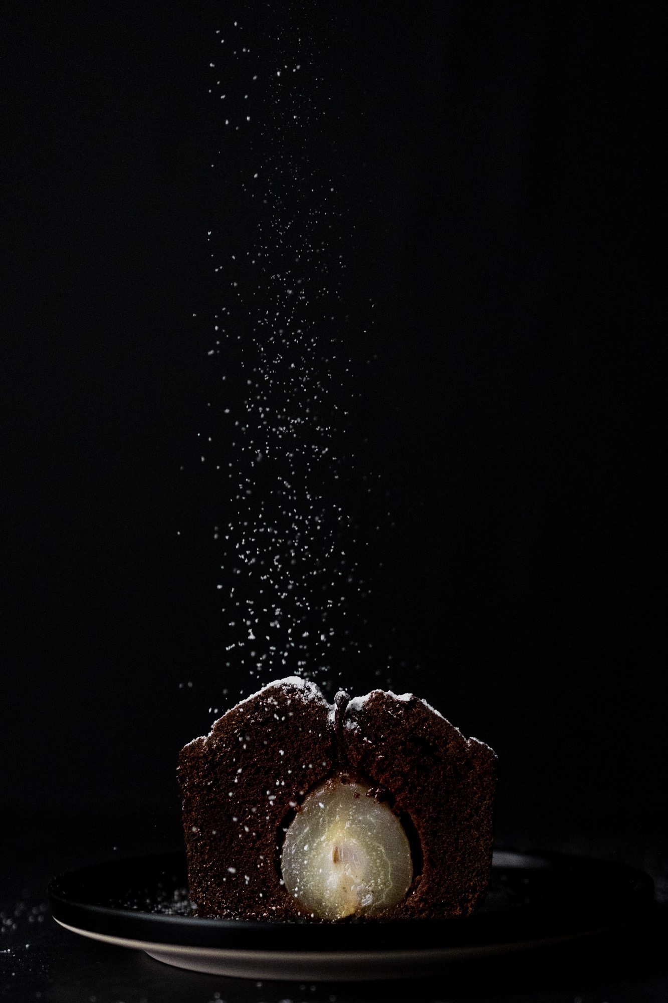 Front view of a chocolate pear cake against black background. The whole pear is inside the cake, creating a bright contrast against the dark backdrop. Icing sugar is falling like snow. Black and white contrast.