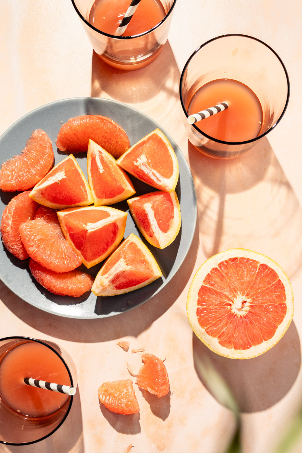 Pieces of juicy orange pink grapefruit on a grey plate, and three glasses with grapefruit juice and straw in a bright summer vibe.