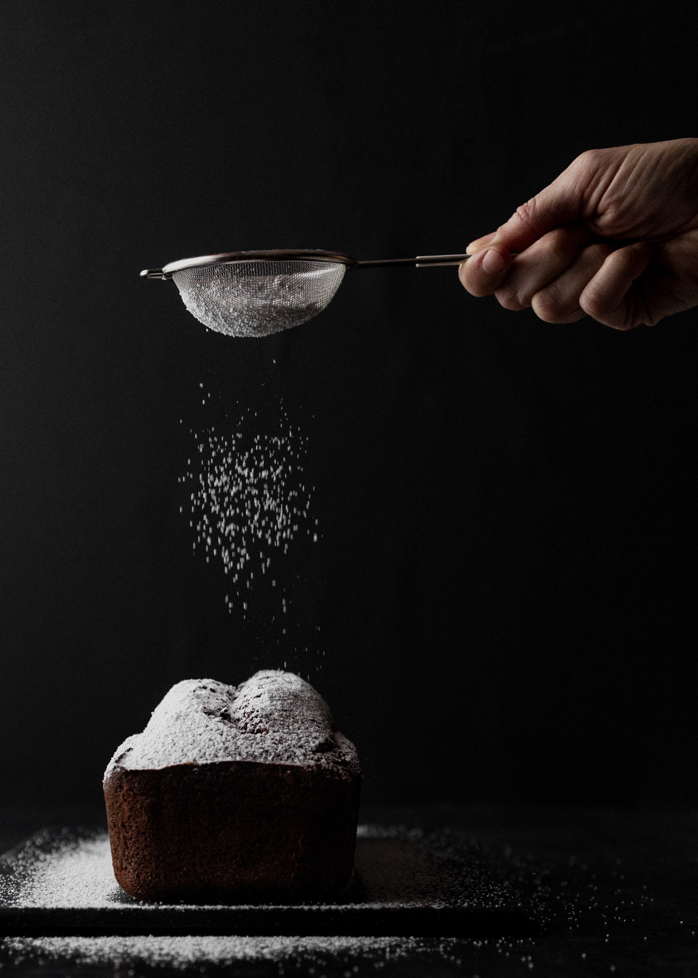 Hand with sieve dusting icing sugar on a cake. The black background creates a high contrast with the white icing sugar.