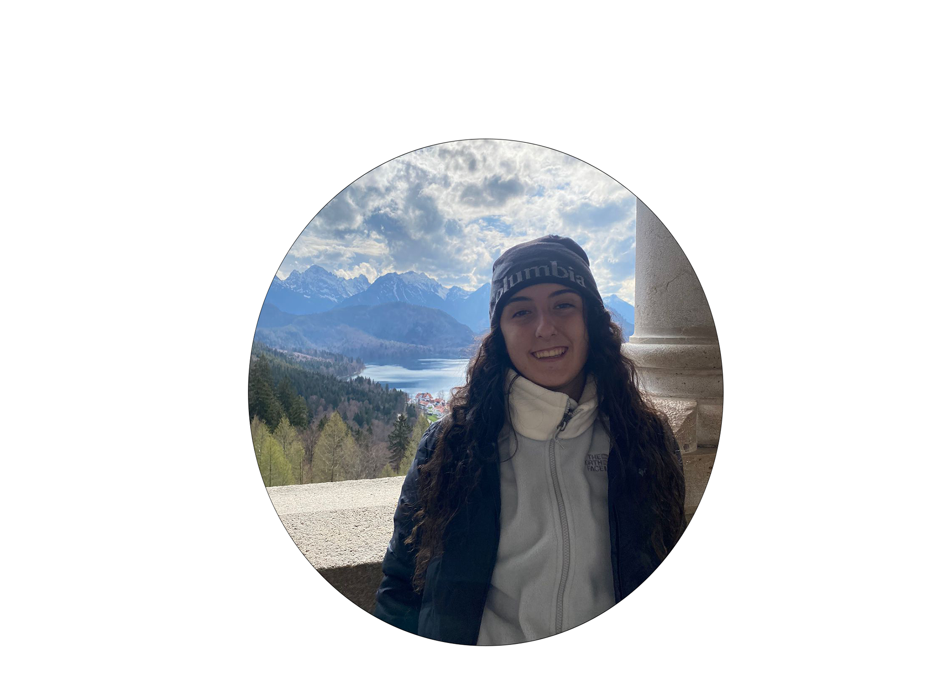 Helenna, standing in a balcony in front of a lake in Germany. She is wearing a white and grey fleece, black winter jacket and black beanie.