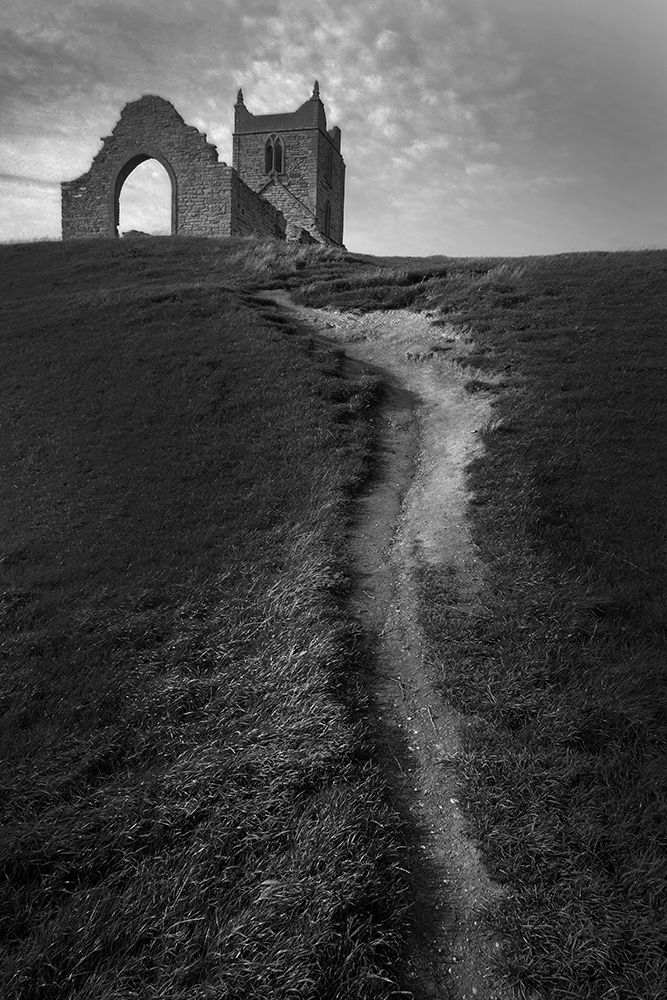 Black and white photograph of. path leading through a field to the ruins of the church called Burrow Mump in Somerset, England