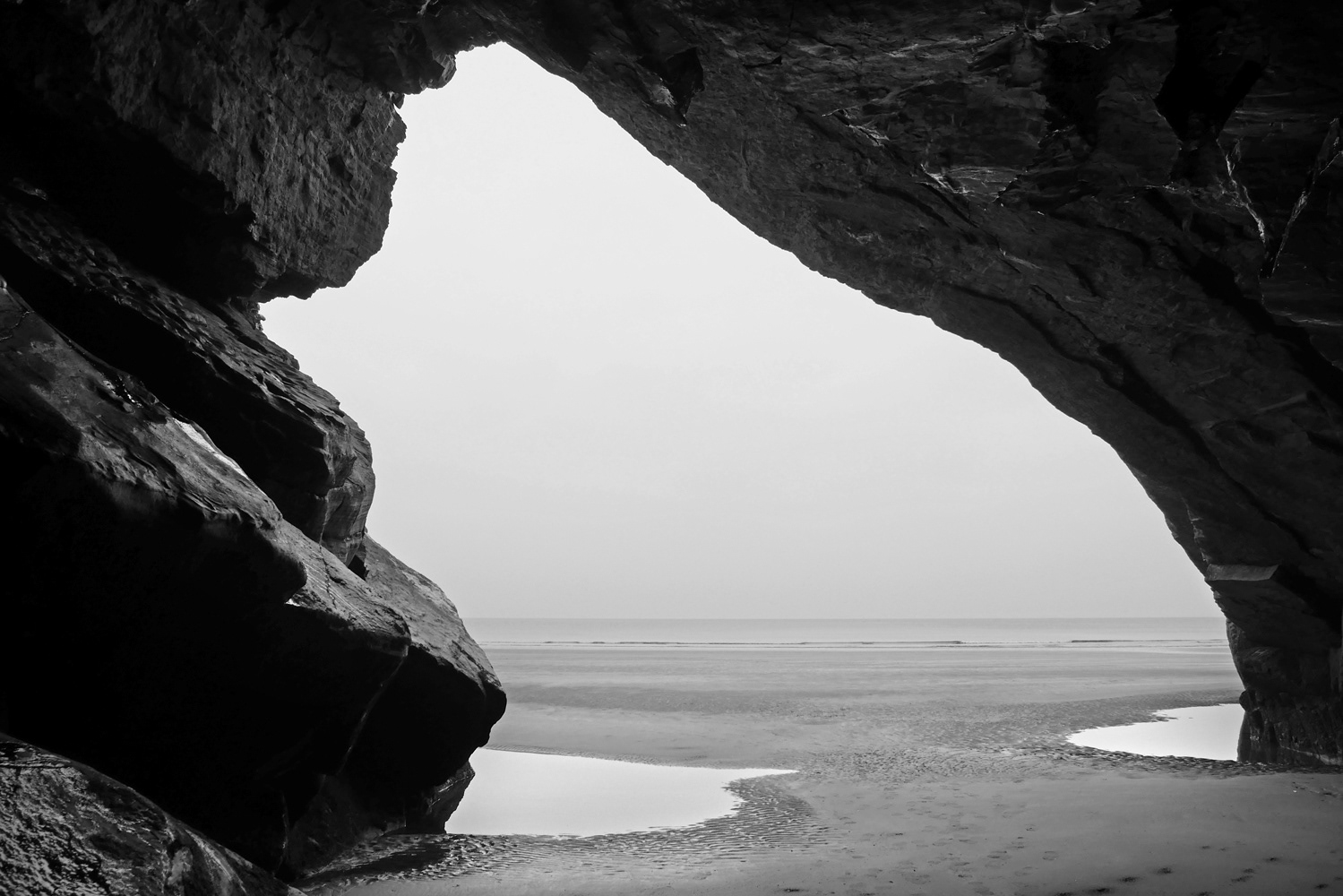 A black and white photograph looking out of black rock cave towards the sea. Wales, UK.