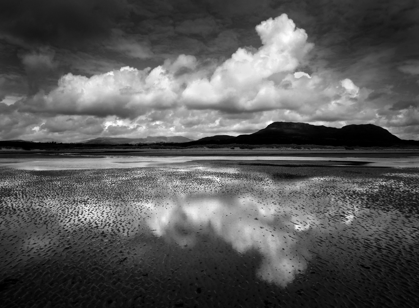 Black and white beach photo showing reflected clouds on beach at low tide.