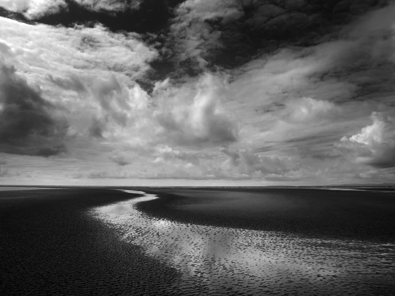Black and white beach photograph of a pool of water leading out to sea with dramatic clouds.